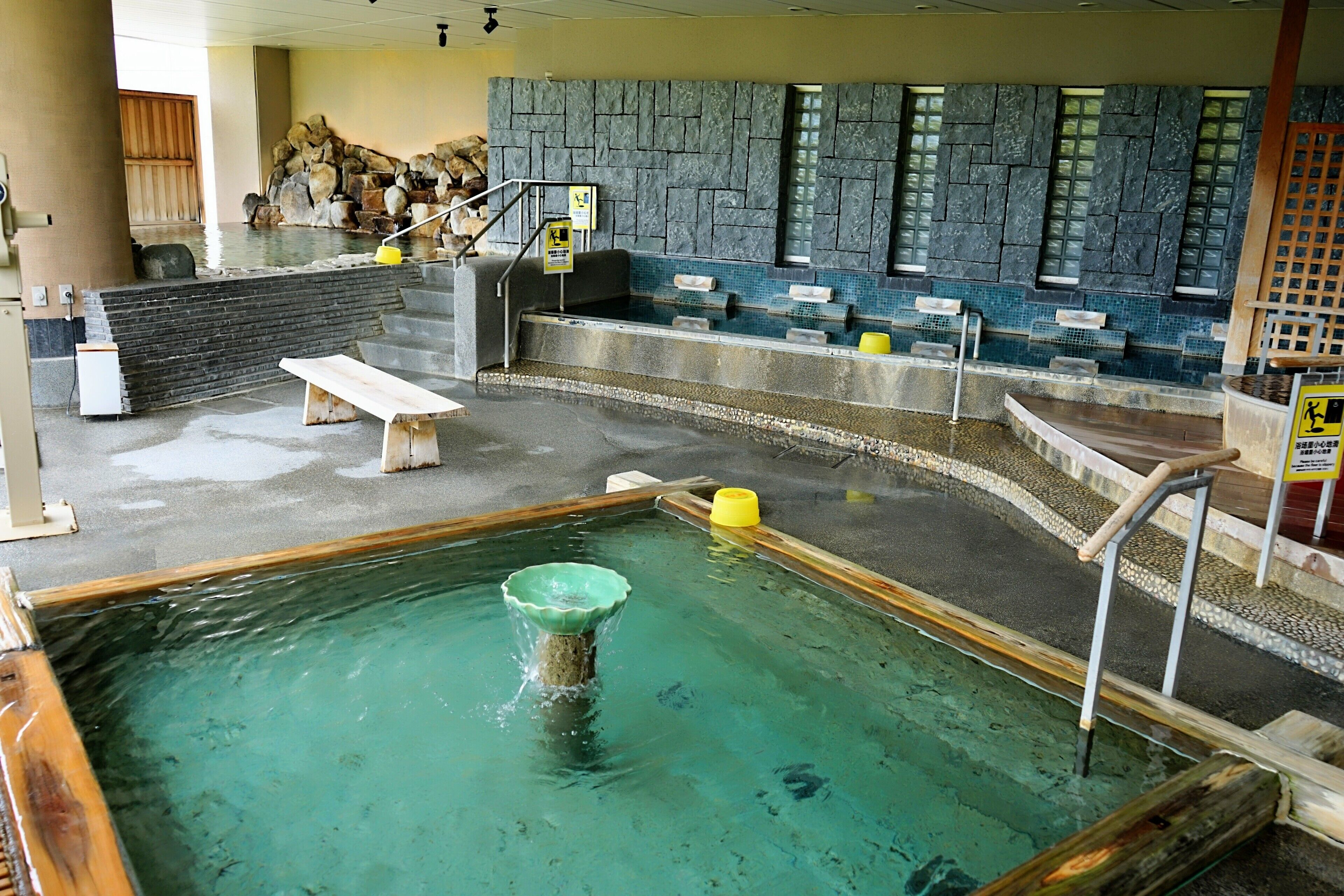A hot spring bath area with stone walls, a wooden bench, and a small pool with a fountain.
