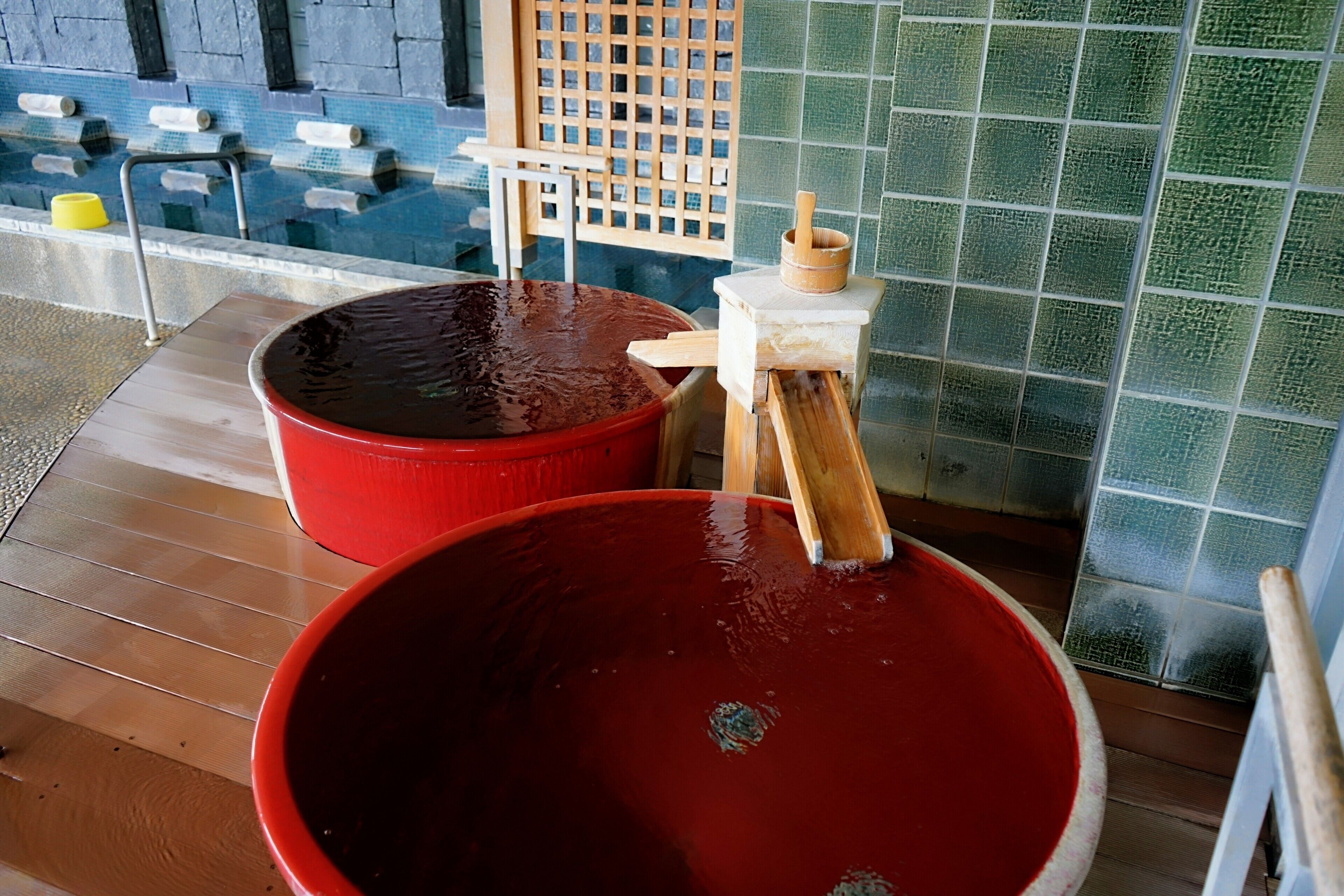 A spa area with a large red basin and a smaller wooden tub, a wooden lattice structure, and a tiled wall.