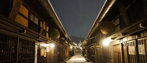 A snow-covered pathway flanked by traditional wooden buildings with illuminated signs, leading towards a distant structure.