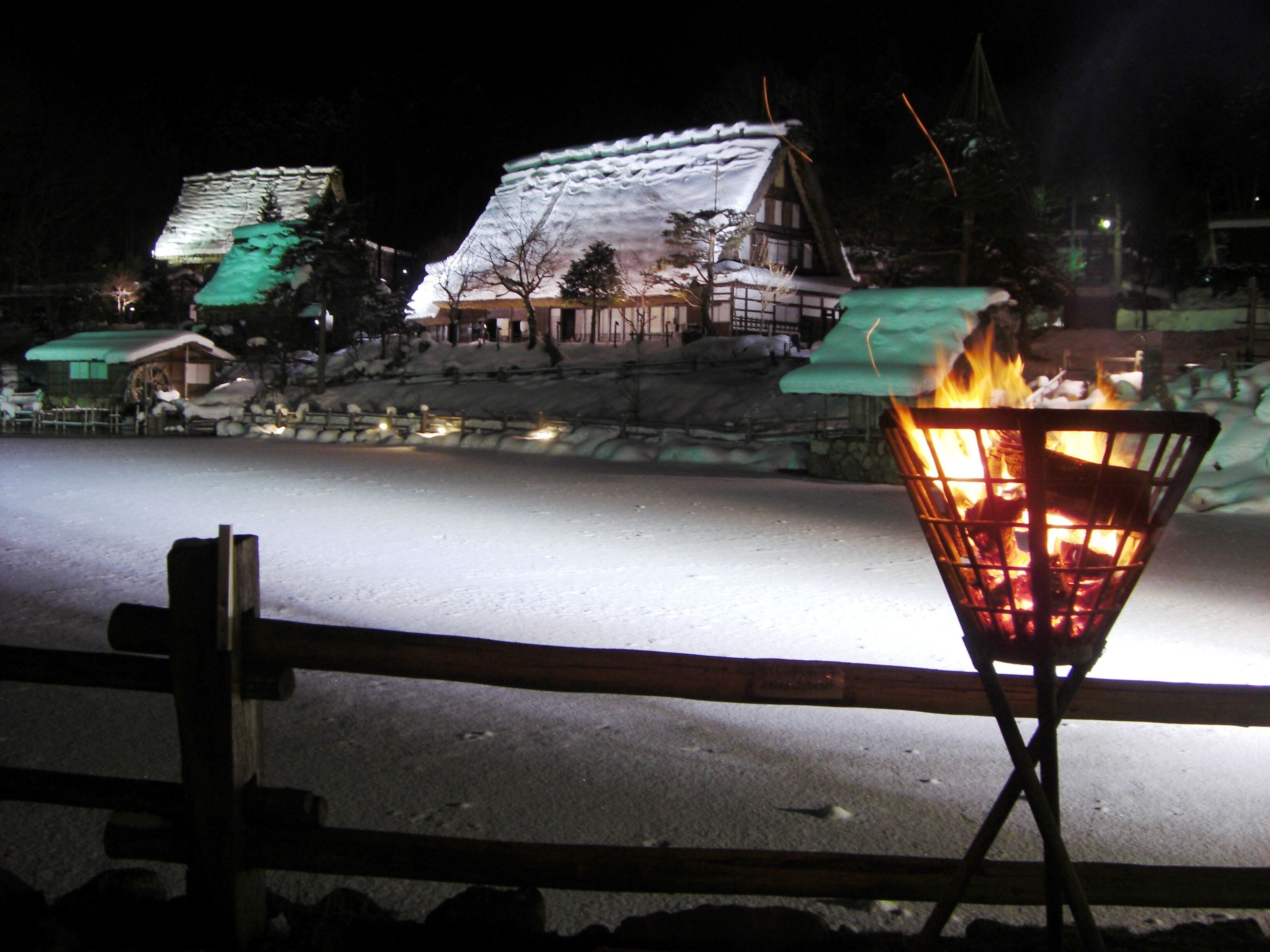 A snow-covered street with a traditional building in the background and a fire in a metal container in the foreground.