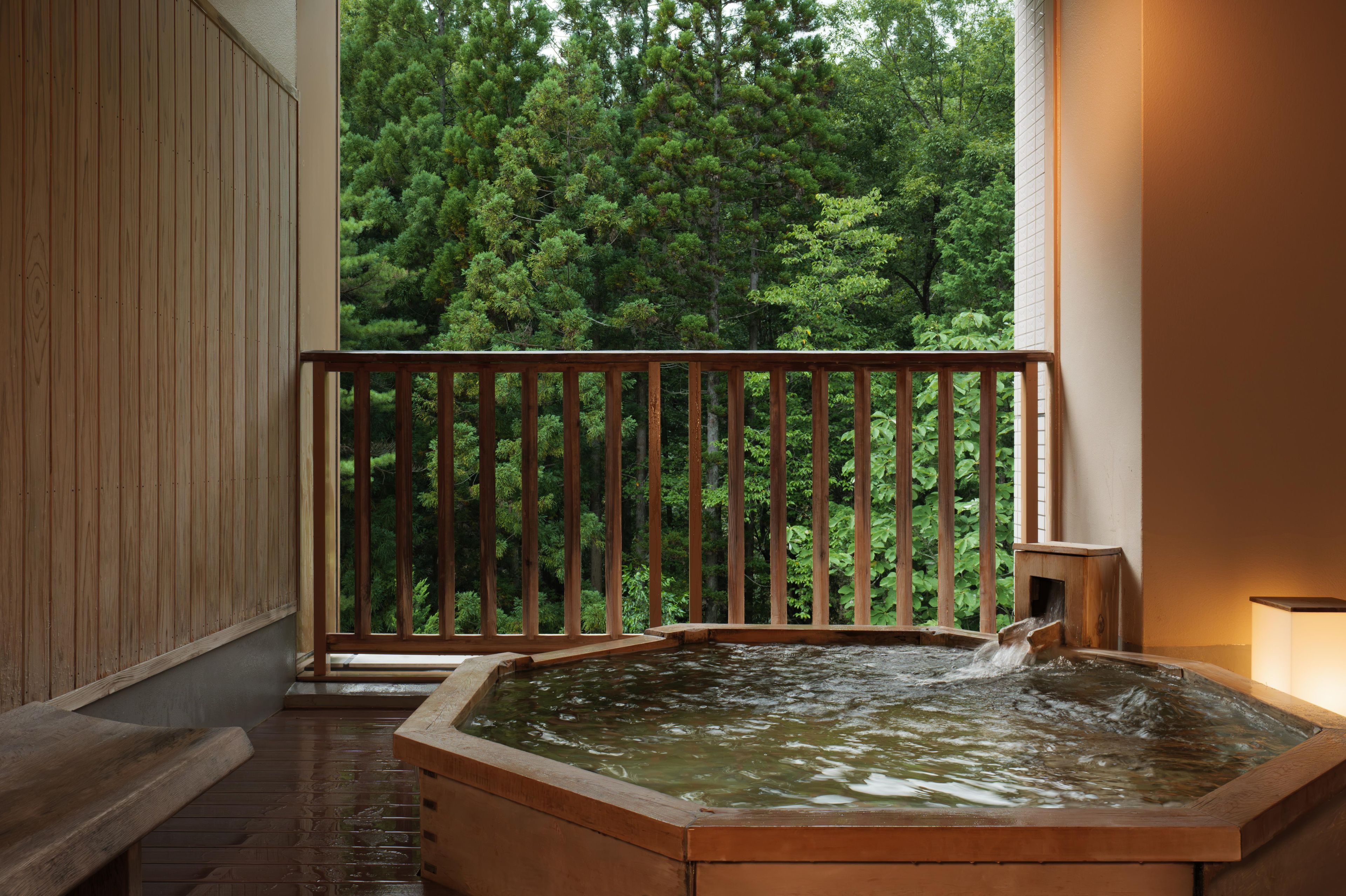 A wooden hot tub with a wooden deck and a railing, surrounded by a forest view.