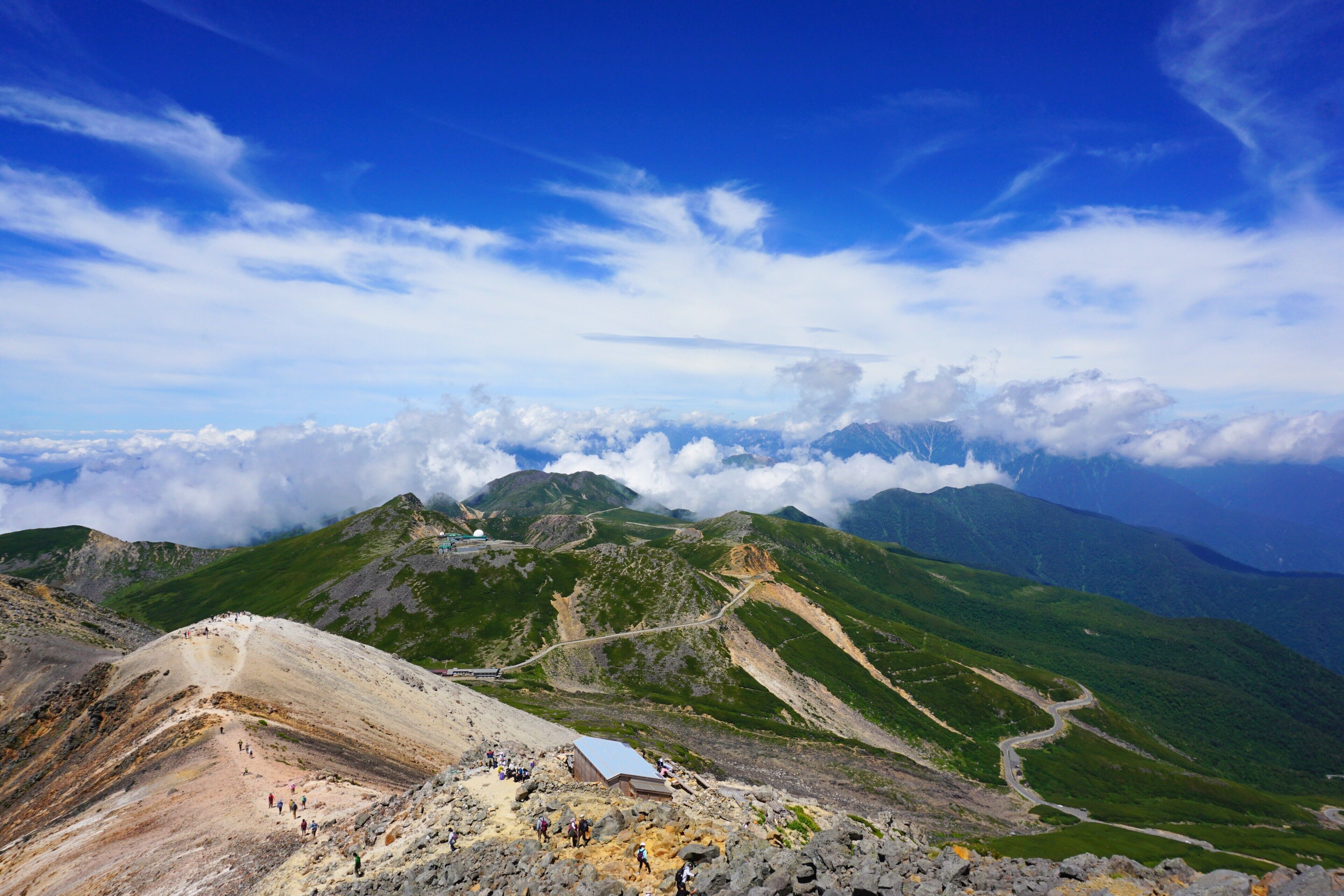 A mountainous landscape with a clear sky, a winding road, and a small building with people around it.