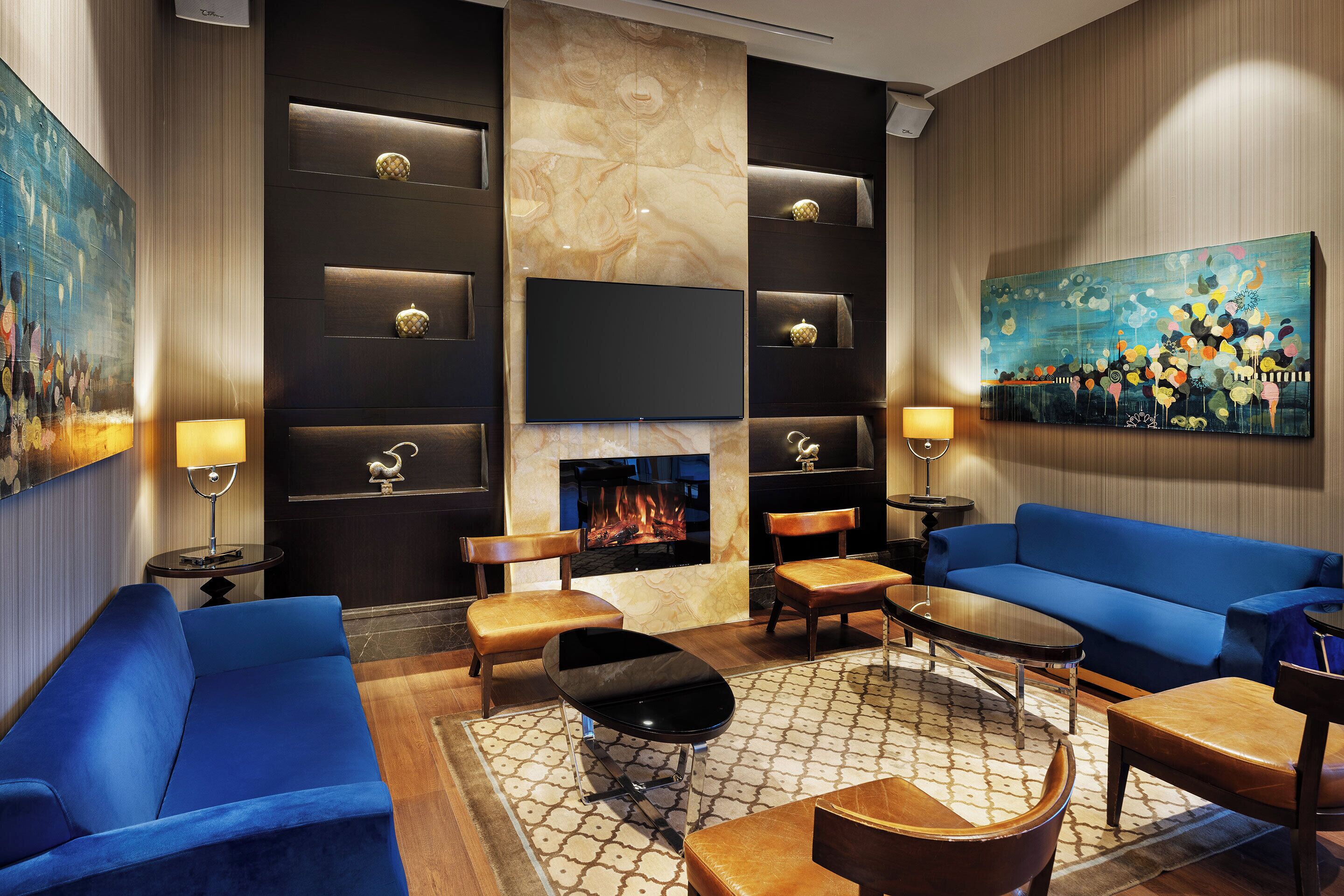 A modern lounge with blue sofas, a flat-screen TV, a fireplace, and a painting on the wall.