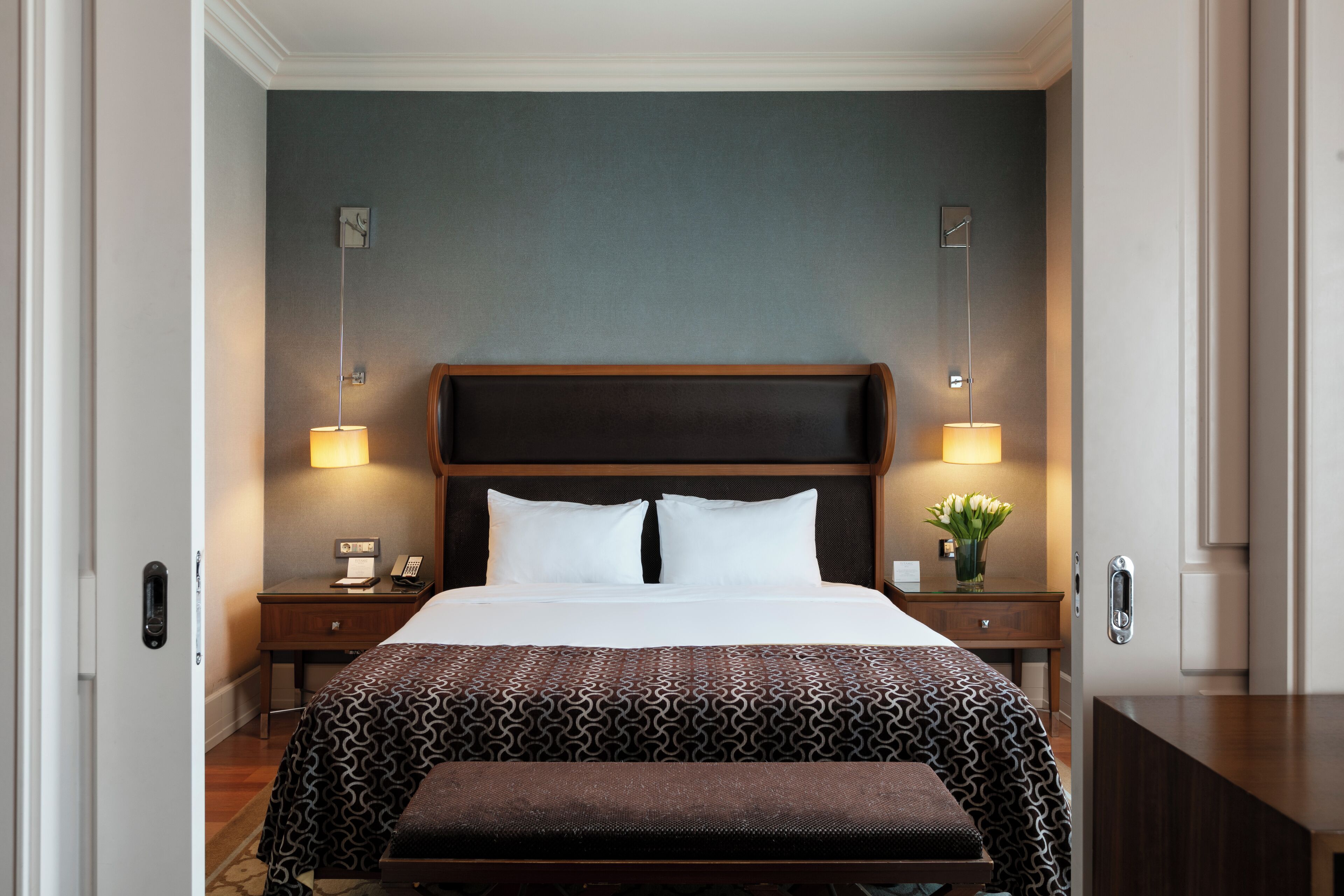 A hotel room with a large bed, two bedside tables with lamps, a nightstand, a bench, and a wall-mounted clock.