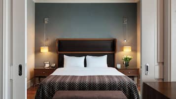A hotel room with a large bed, two bedside tables with lamps, a nightstand, a bench, and a wall-mounted clock.
