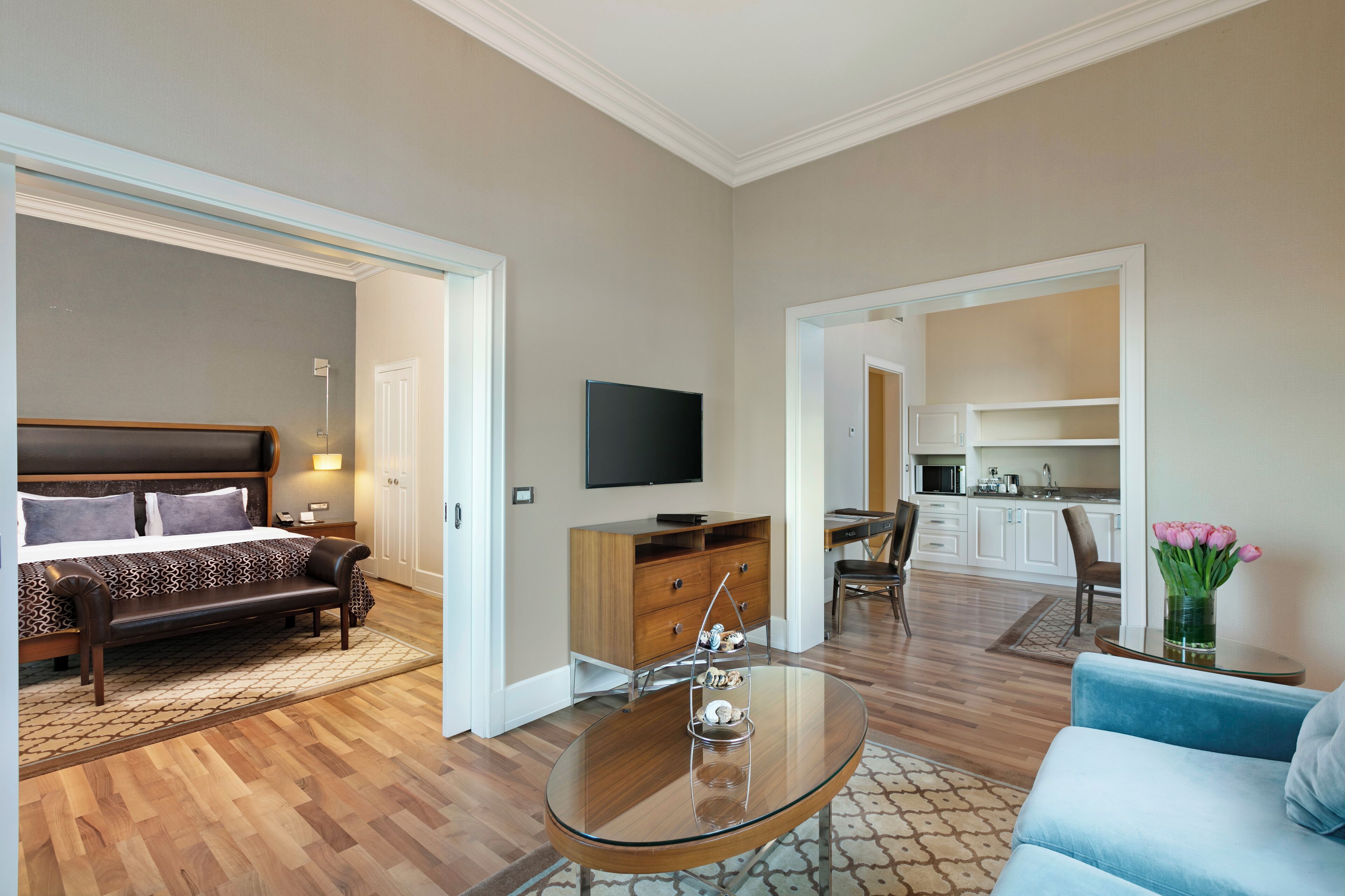 A modern hotel room with a bed, a sofa, a glass coffee table, a TV, a kitchen area, and a bathroom visible through an open door.