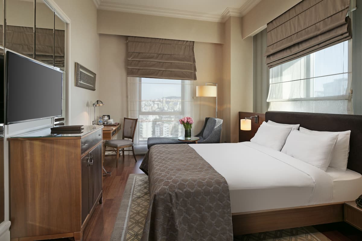 A hotel room with a large bed, a desk with a chair, a TV, and a window with a view of the city.