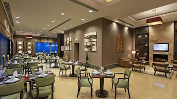 Un amplio vestíbulo de hotel con varias mesas de comedor, asientos cómodos, una chimenea y una pantalla de televisión.
