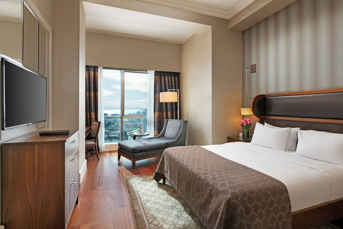 A hotel room with a bed, a desk, a chair, a TV, and a view of the city.