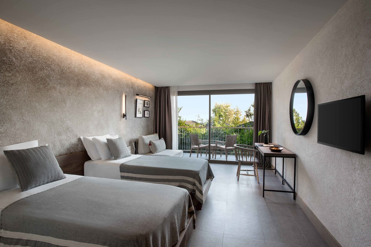 A hotel room with two beds, a TV, a balcony with a table and chairs, and a view of the outdoors.