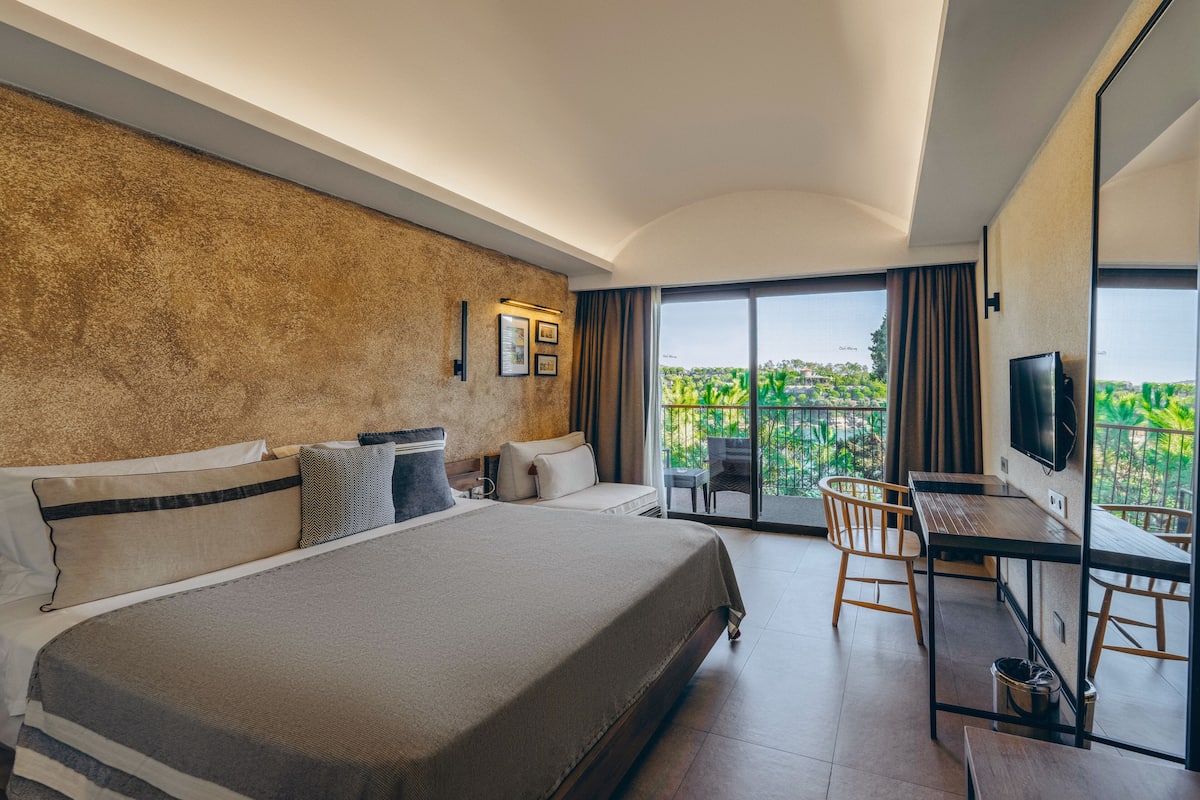 A hotel room with a large bed, a desk, and a TV. There is a balcony with a view of greenery.