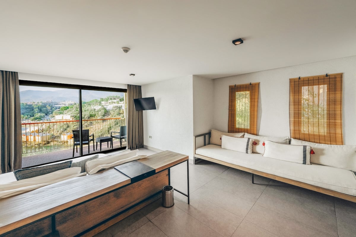 A modern hotel room with a large balcony, a sofa, a wooden table, and a TV mounted on the wall.