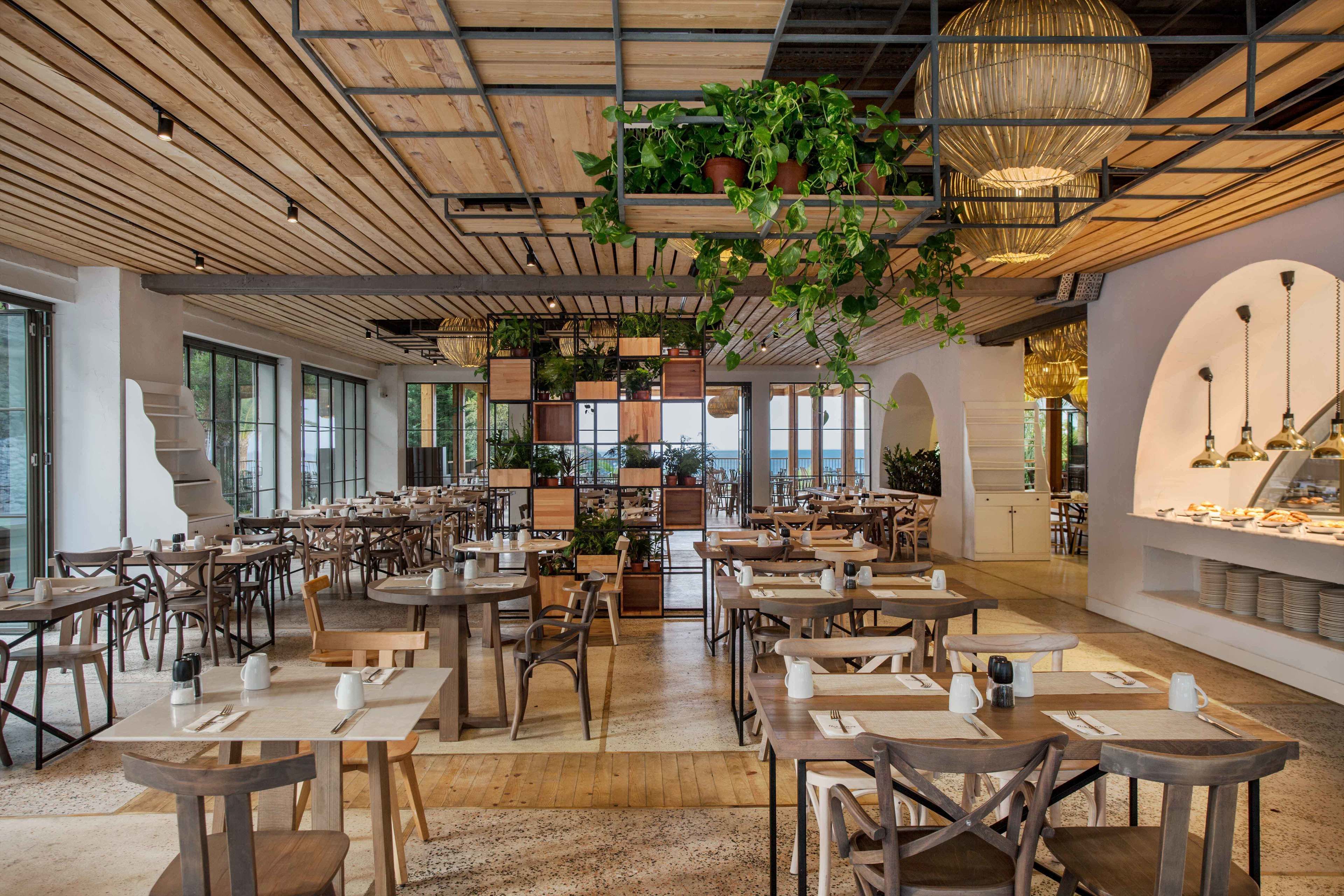Ein geräumiges Restaurant mit Holztischen und -stühlen, verschiedenen Pflanzen und einer modernen Kronleuchter.
