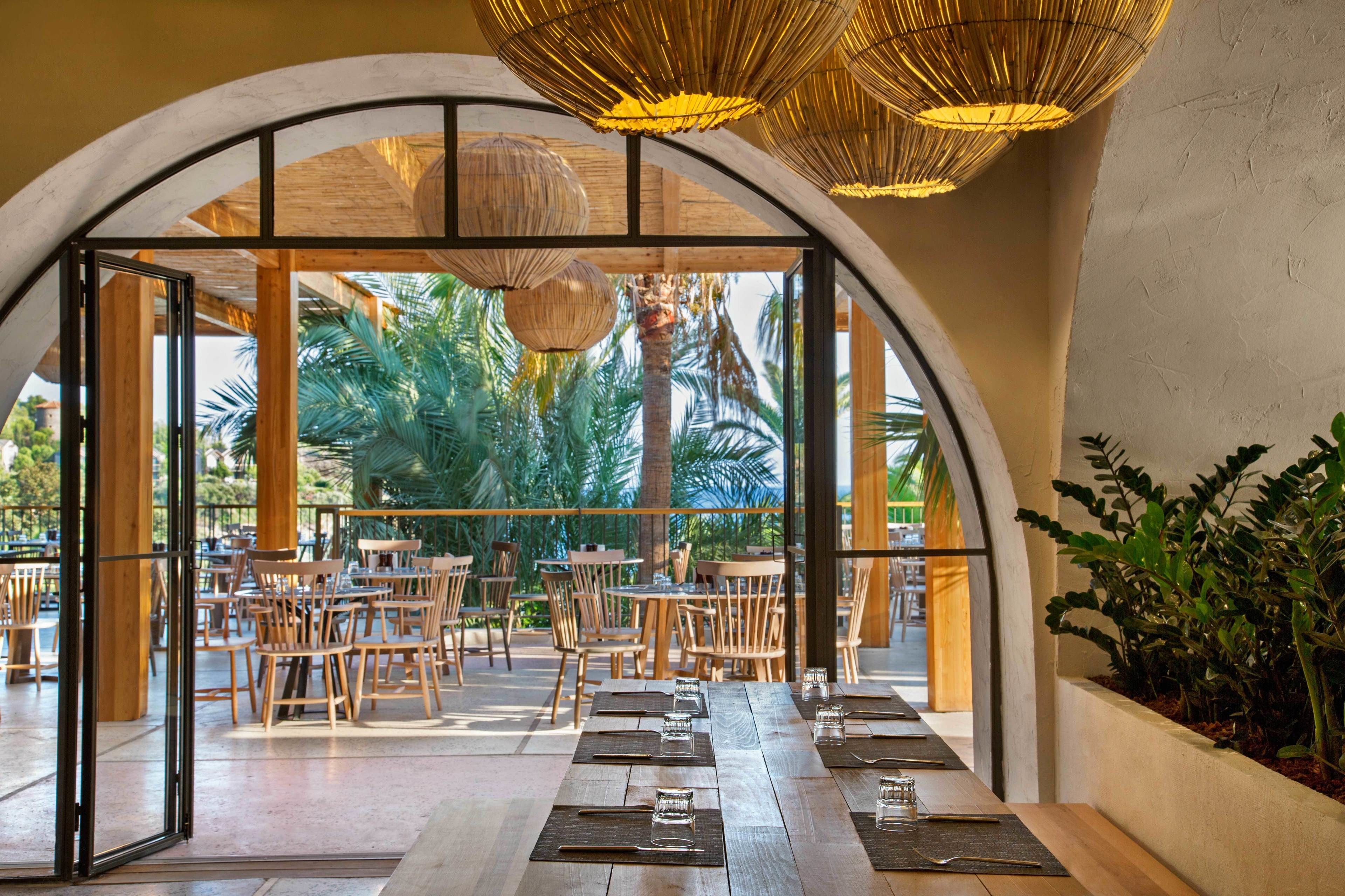 An indoor dining area with wooden tables and chairs, large windows, and hanging wicker lamps.