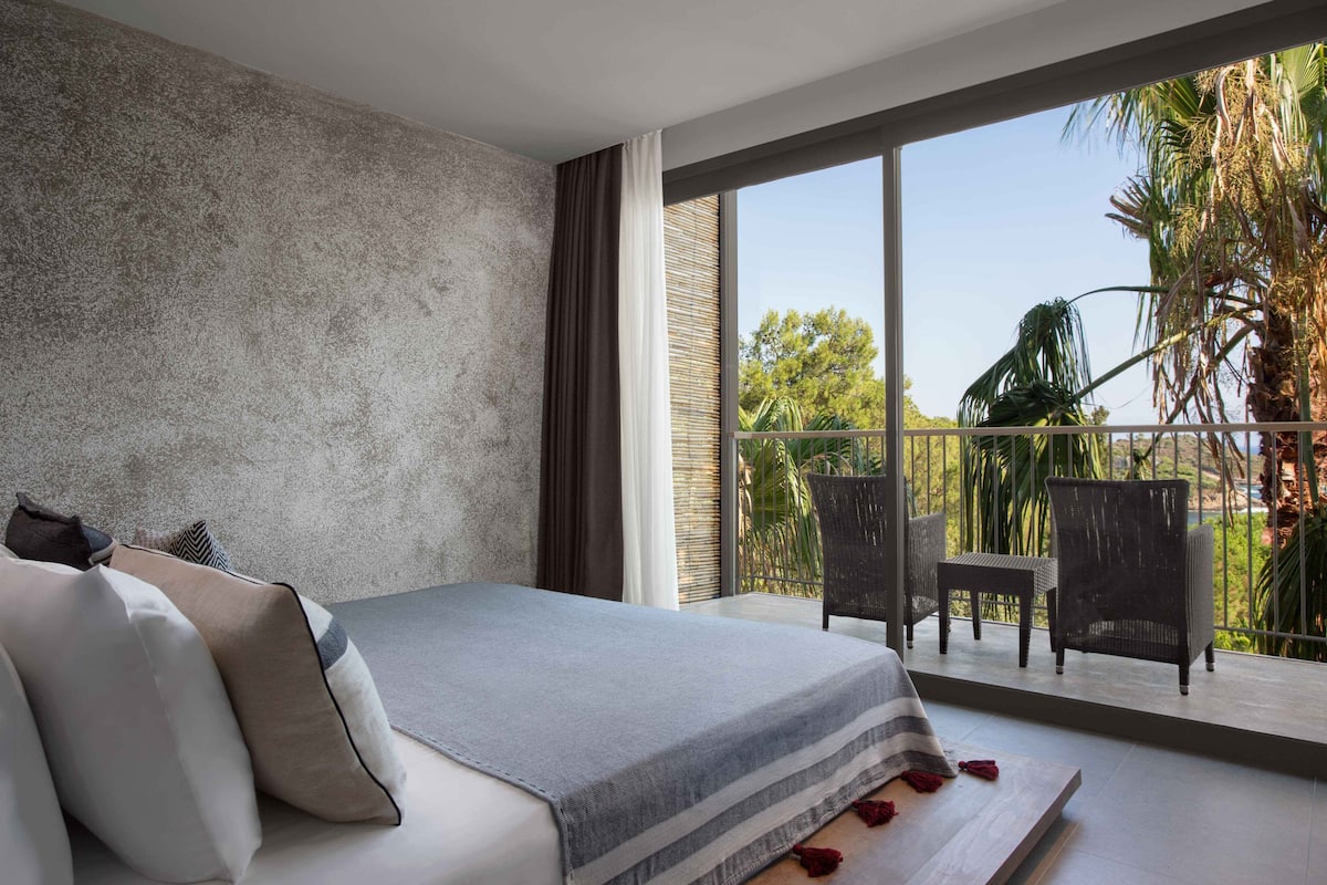A bedroom with a large bed, a balcony with chairs, and a view of palm trees.