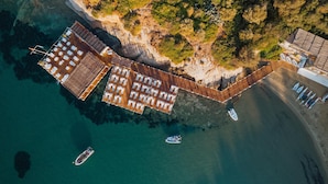 Vista aérea de un complejo turístico costero con muelle de madera, numerosas tumbonas y un pequeño barco atracado cerca de la orilla.
