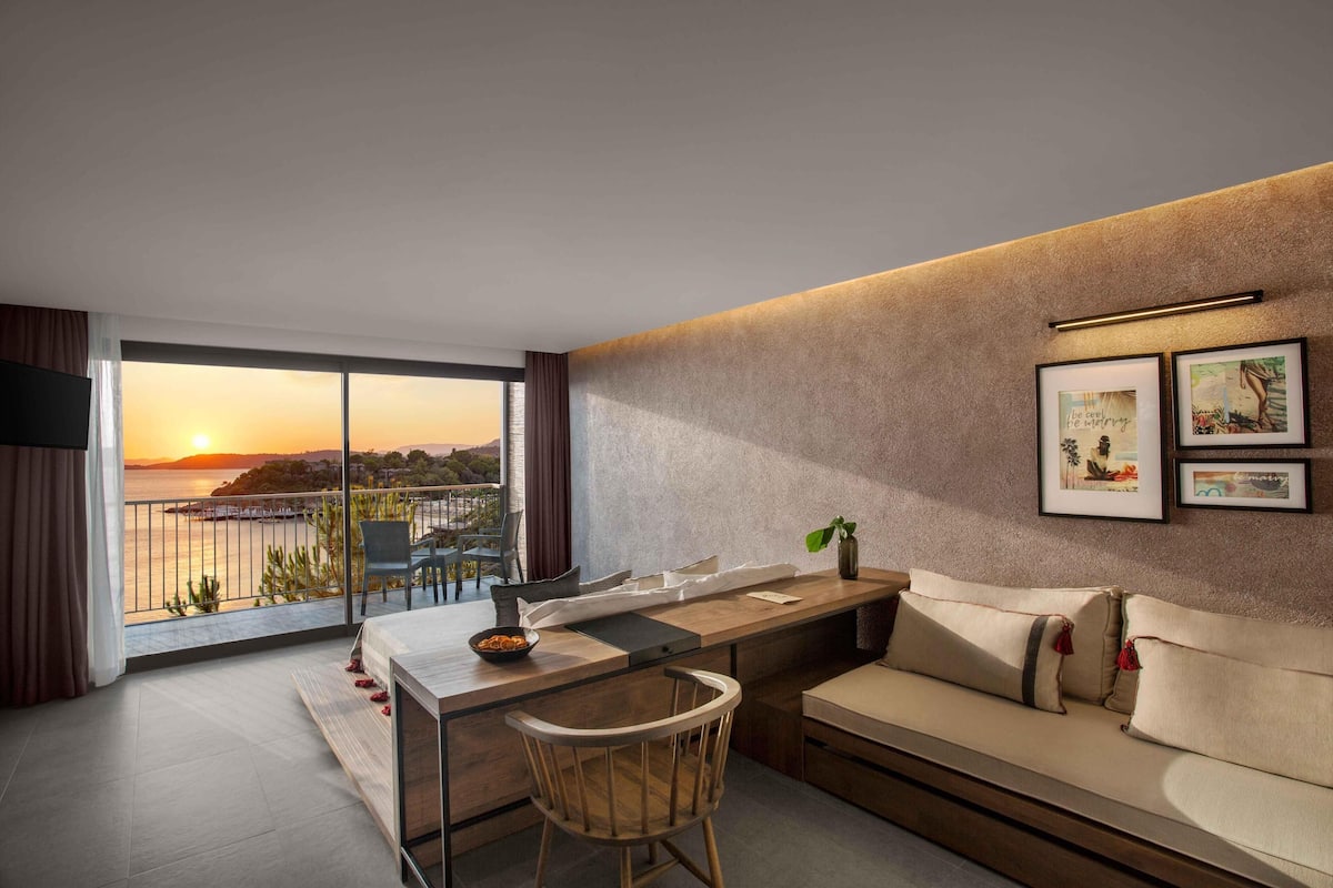 A modern living room with a sofa, a dining table, and a view of the sunset through large windows.