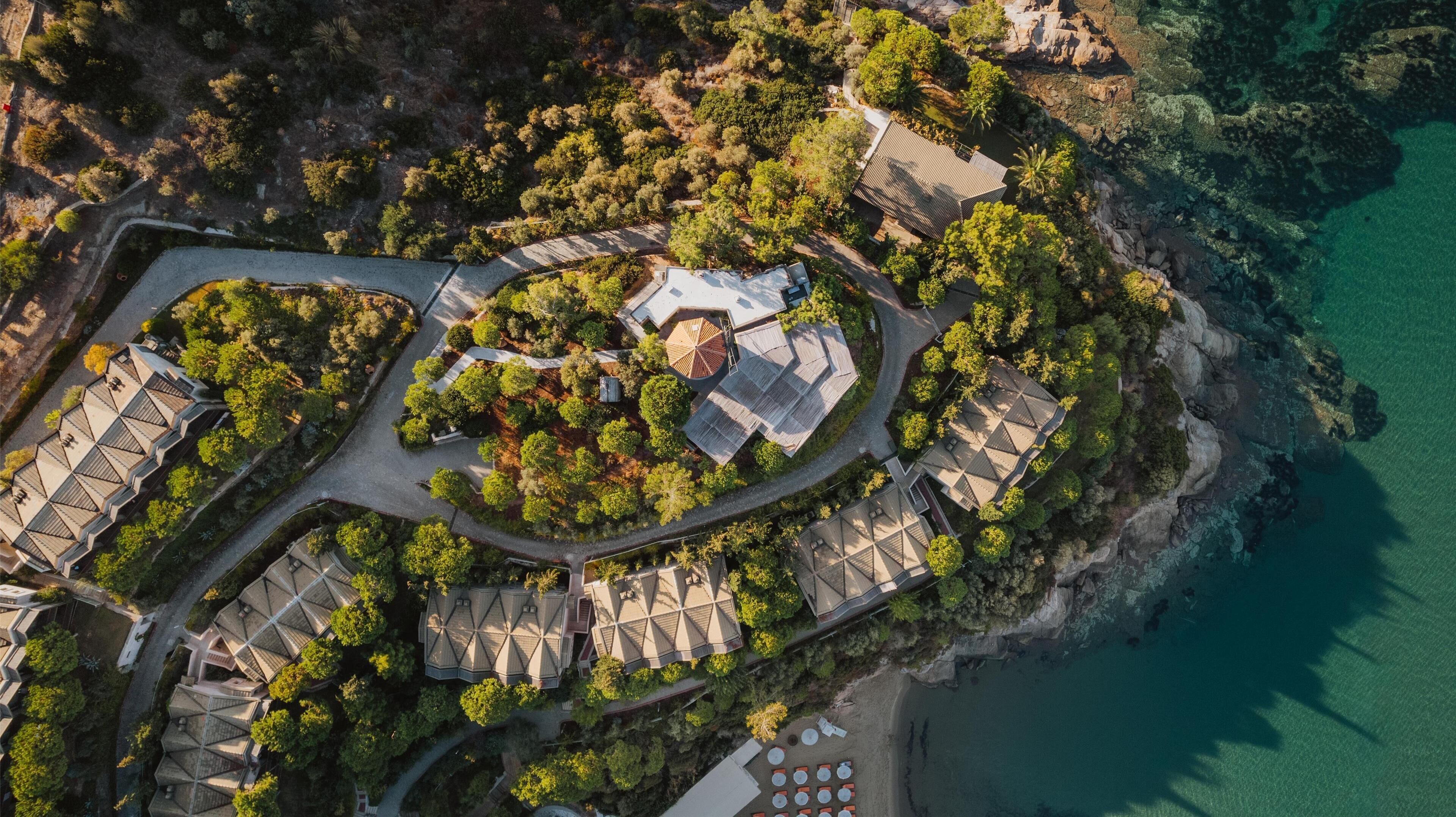 Luftaufnahme eines Küstenresorts mit mehreren Gebäuden, einer zentralen, kreisförmigen Struktur und einer umliegenden, bepflanzten Anlage.