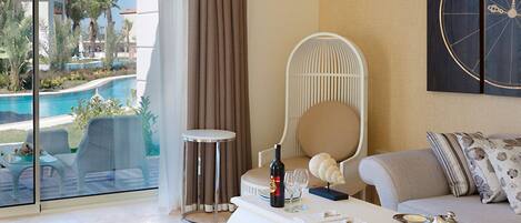 A modern hotel room with a glass door leading to a pool area, a white curved coffee table with a bottle and glasses, a white chair, and a sofa with decorative pillows.