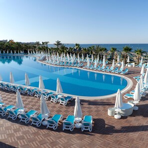A large swimming pool with blue water, surrounded by rows of white lounge chairs and umbrellas, and palm trees in the background.