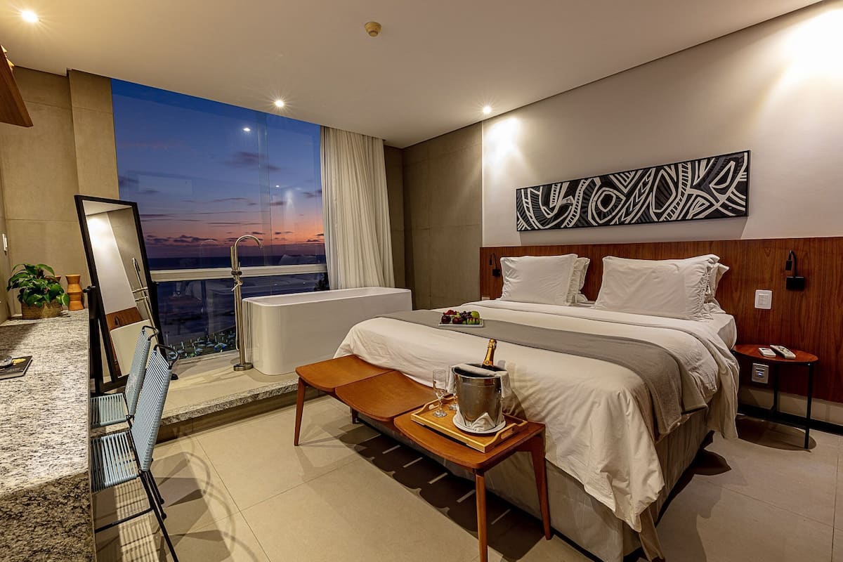A modern hotel room with a large bed, a bathtub, a dining table, and a city view.