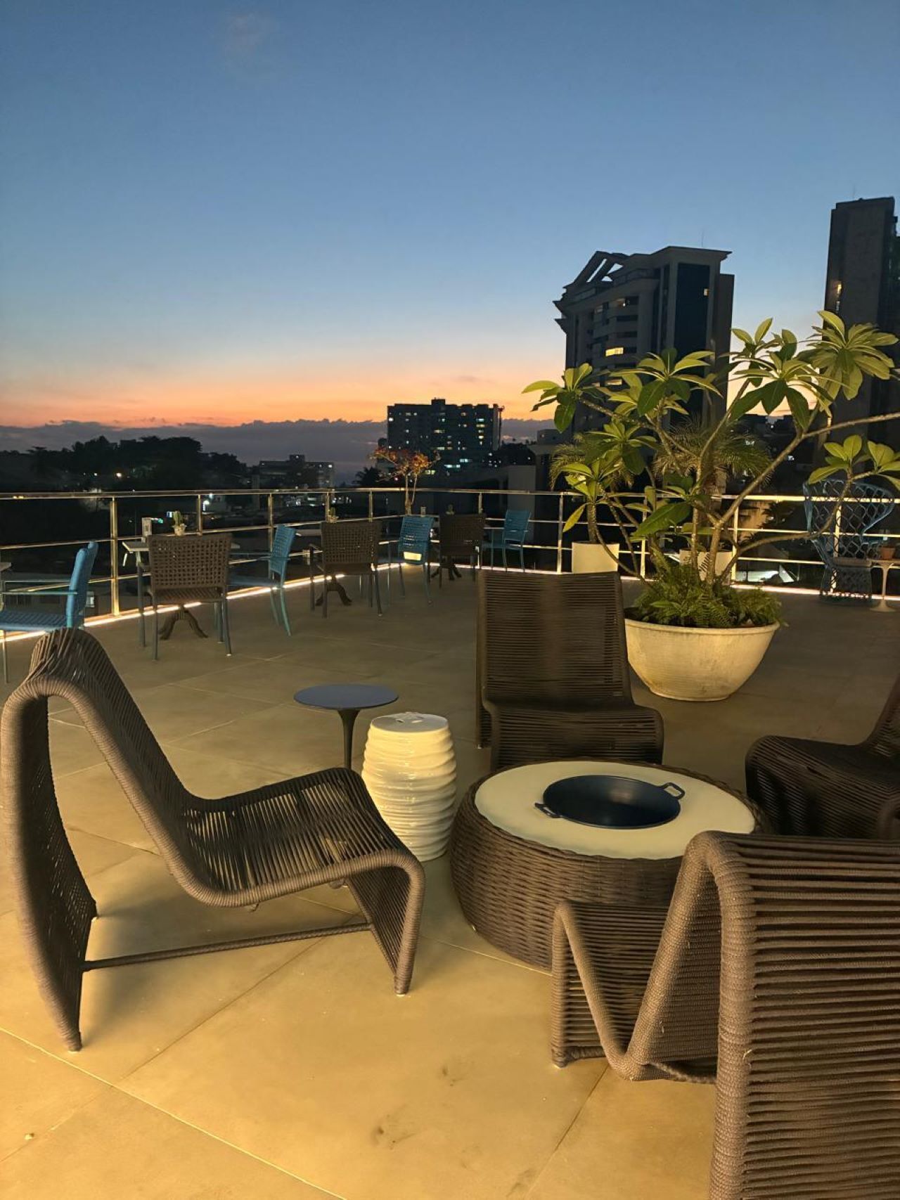 Terraza en la azotea con mobiliario de mimbre, una planta en maceta y vistas de edificios de la ciudad al atardecer.