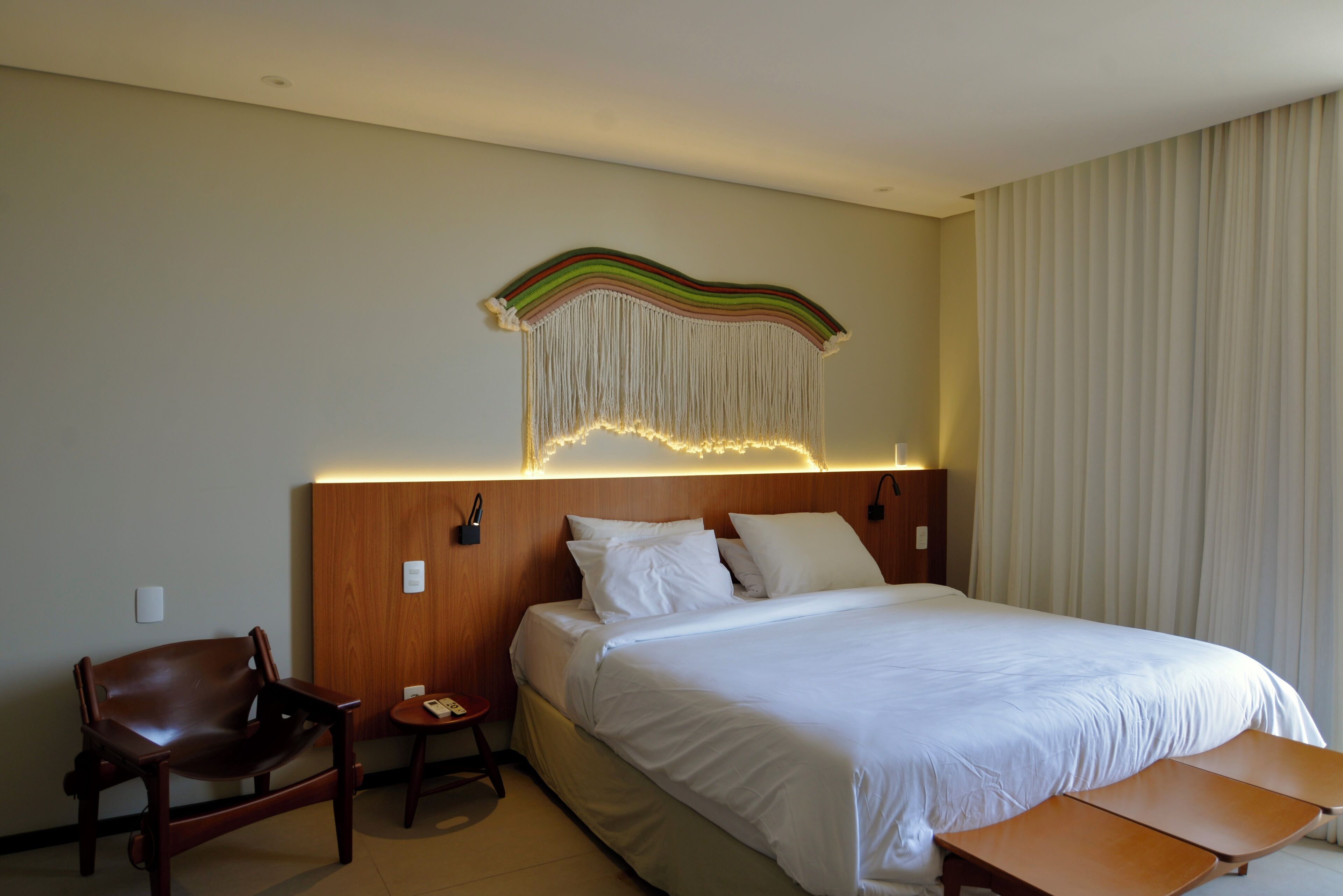 A hotel room with a bed, a wooden headboard, a chair, a small table, and a wall-mounted light fixture.