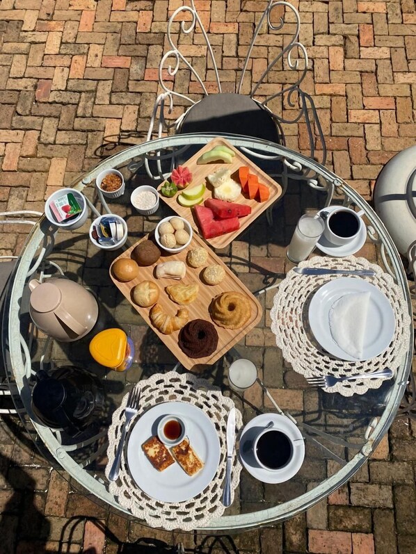 A round glass table with a variety of breakfast items including pastries, fruits, and beverages, set on a brick pavement. - Zank by Toque Hotel (Salvador)
