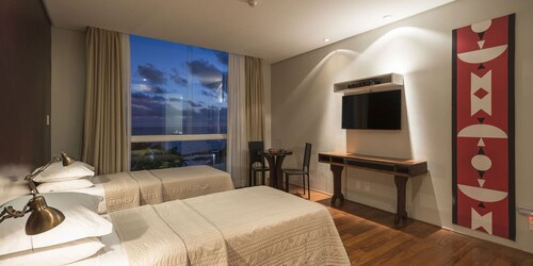 A hotel room with two beds, a desk, a chair, a TV, and a large window with a view.
