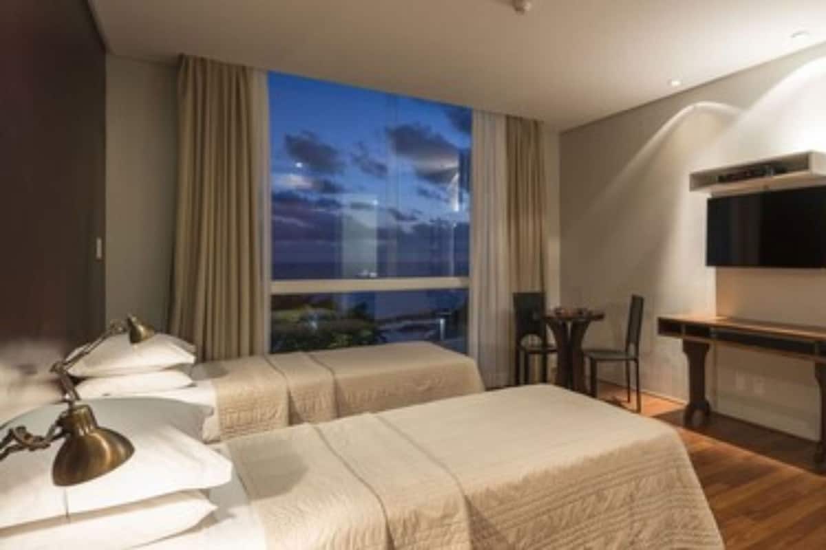 A hotel room with two beds, a desk, a chair, a TV, and a large window with a view.