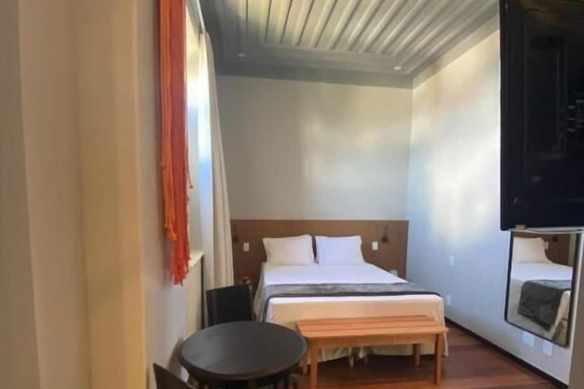 A hotel room with a bed, a small table, a chair, and a TV mounted on the wall.