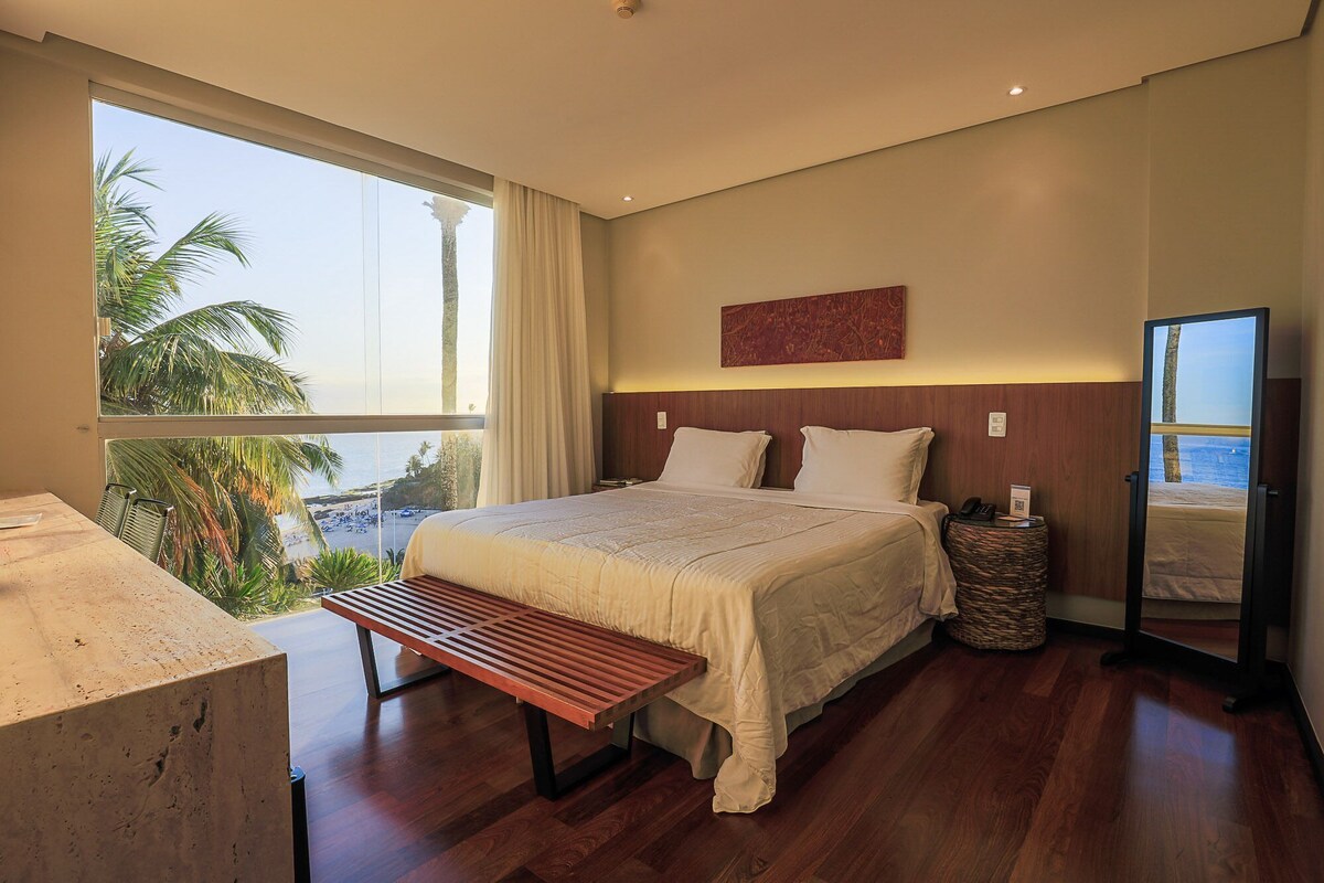 Habitación de hotel con una cama grande, un banco, una mesita de noche, un espejo y vista a la playa.