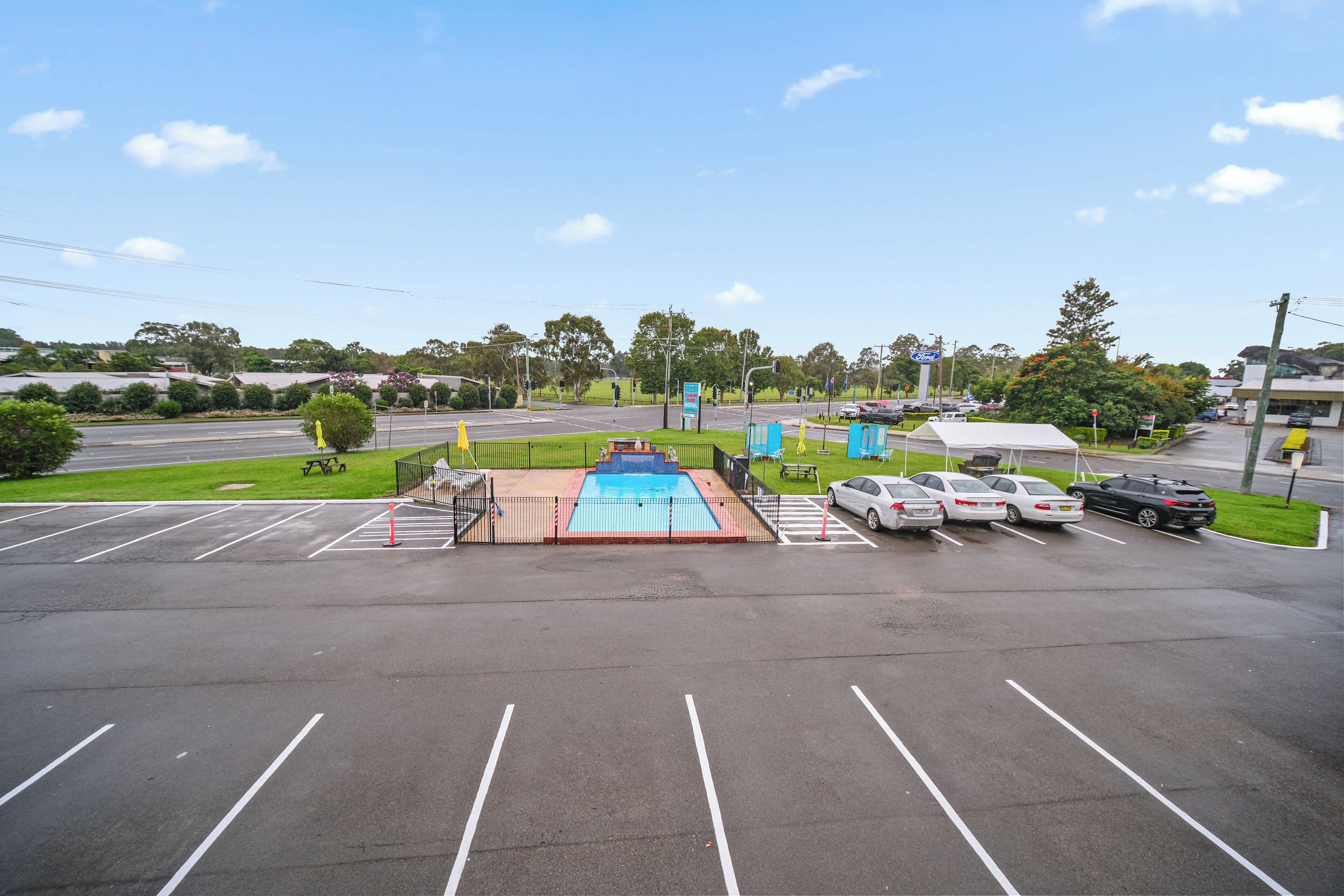 A parking lot with a swimming pool, several cars, and a green lawn area.
