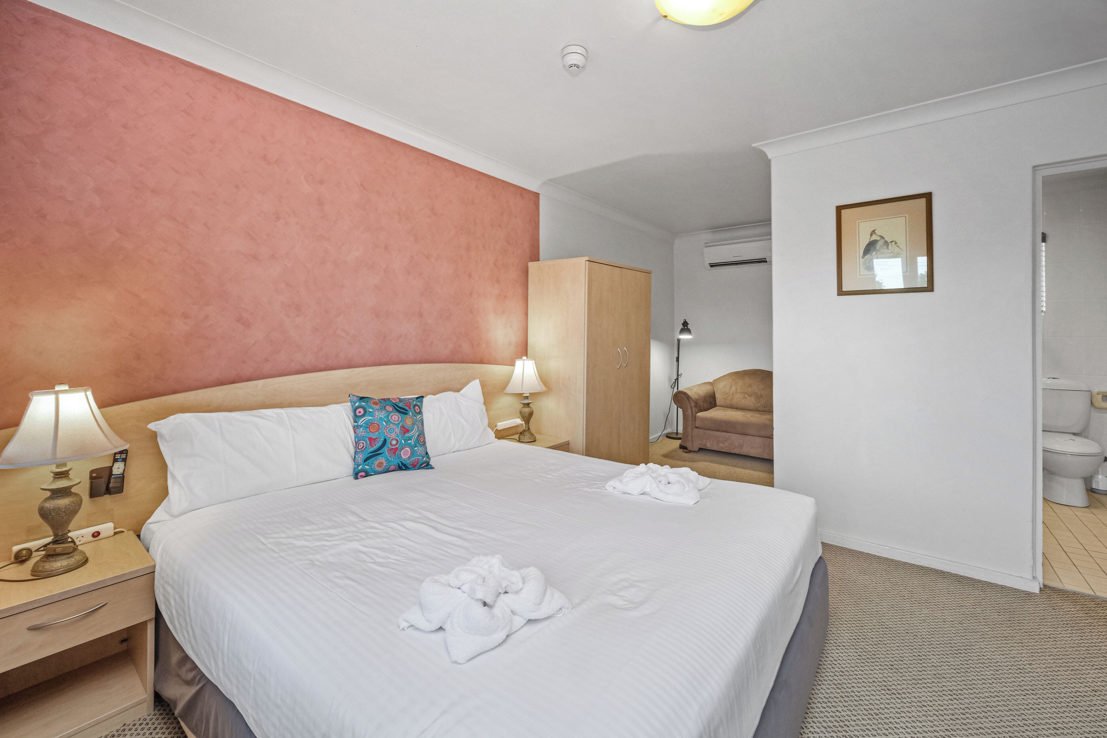 Habitació d'hotel amb un llit gran, un armari, un escriptori amb una làmpada i un bany visible a través d'una porta oberta.