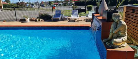 En swimmingpool med en statue, liggestole og en rød og hvid parasol.