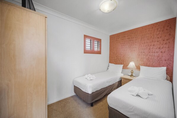 A hotel room with two beds, a wardrobe, a small window, and a nightstand with a lamp.