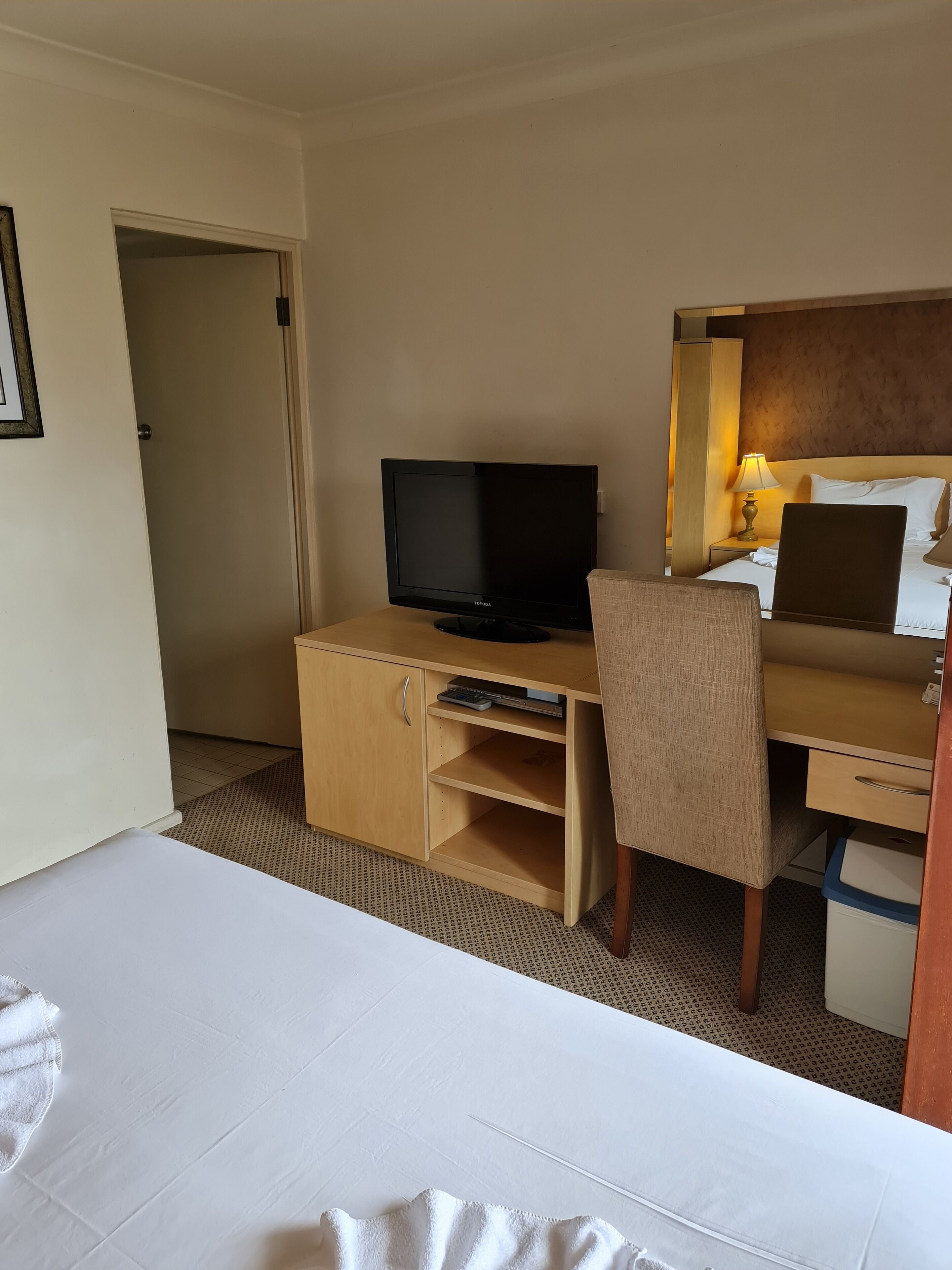 A hotel room with a bed, a desk with a television, a chair, and a bedside table with a lamp.