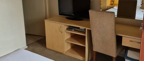 A hotel room with a bed, a desk with a television, a chair, and a bedside table with a lamp.