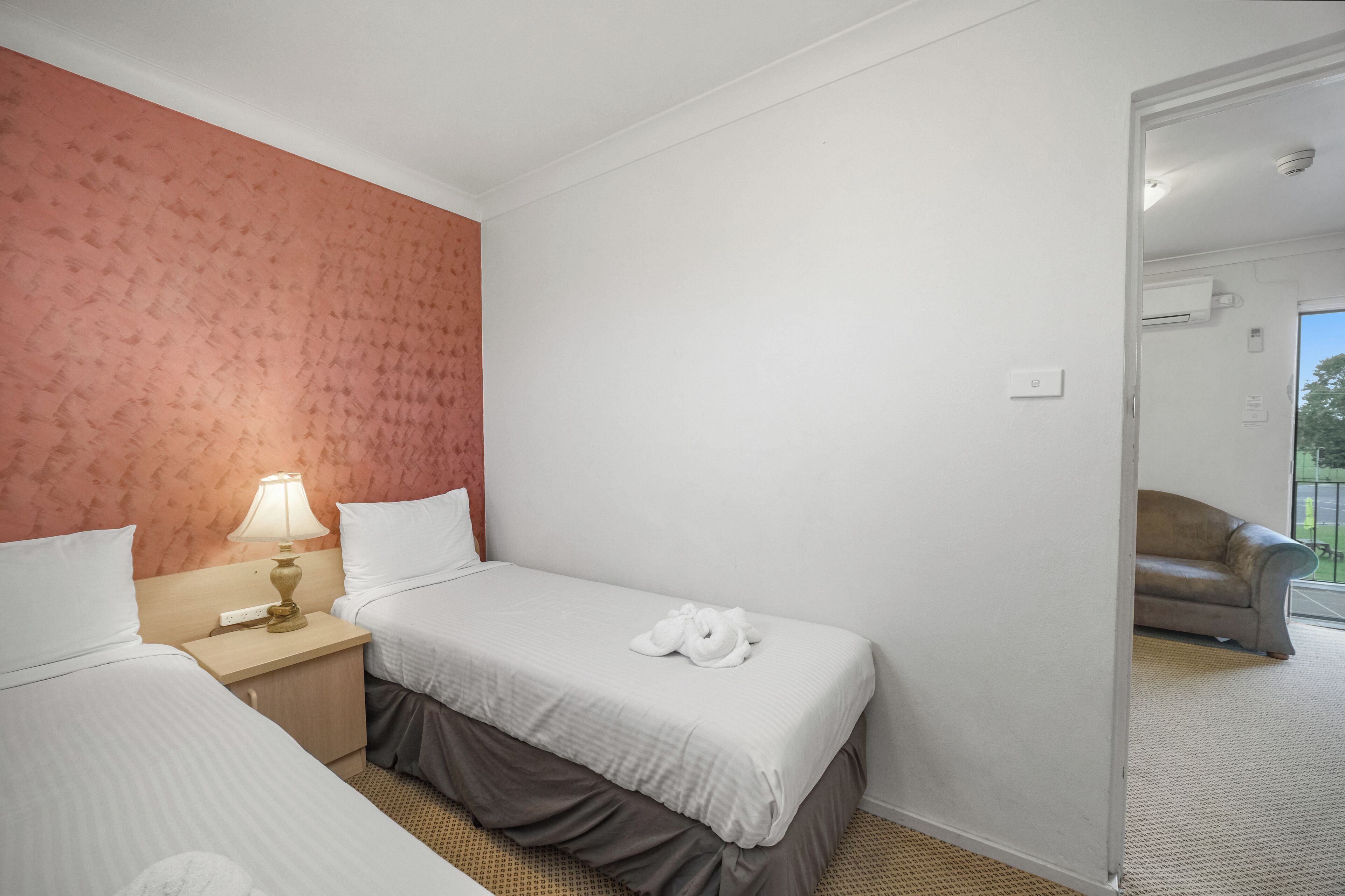 Une chambre d’hôtel avec deux lits, une table de chevet avec une lampe et une vue sur un balcon équipé d’un canapé.