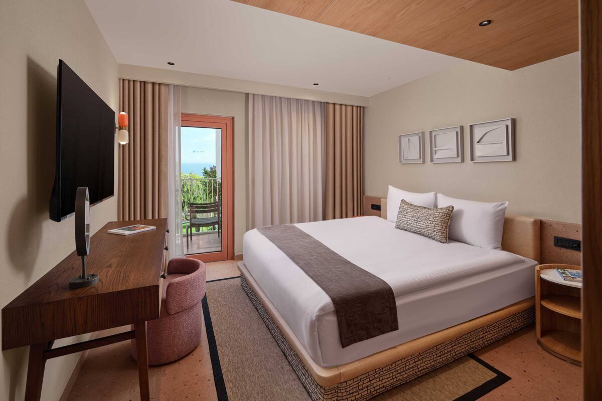 Een hotelkamer met een groot bed, een bureau met stoel, een aan de muur bevestigde televisie en uitzicht op de omgeving via een deur.