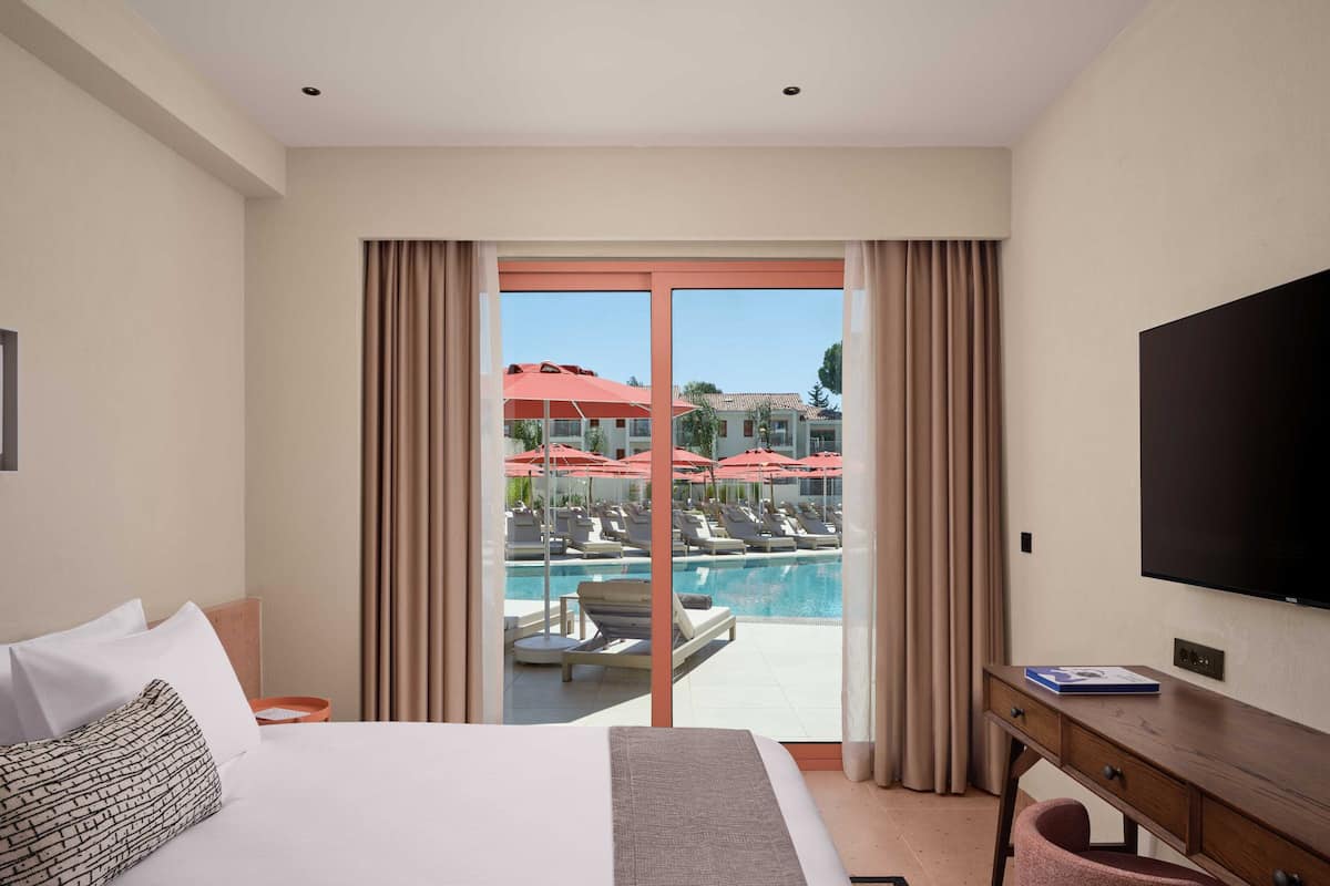 Hotelkamer met een bed, een bureau, een televisie en uitzicht op een zwembad en parasols.