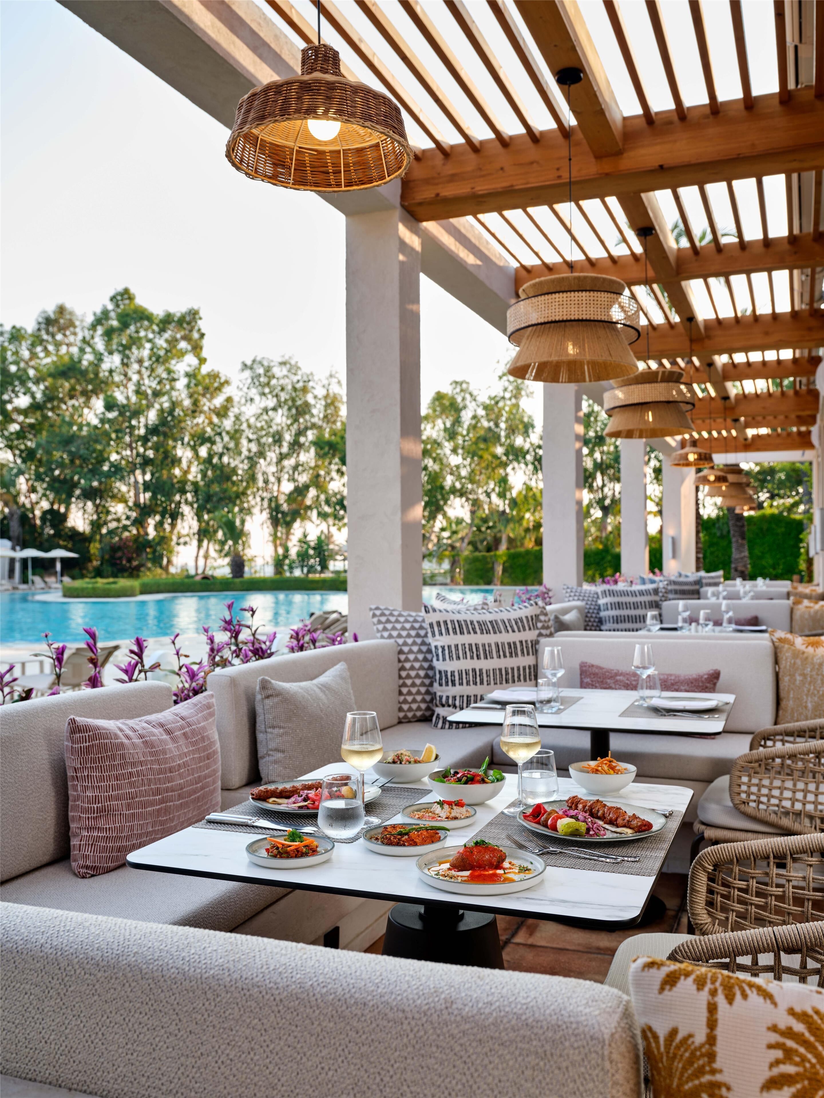 An outdoor dining area with multiple tables and chairs, a pool in the background, and a wooden pergola overhead.