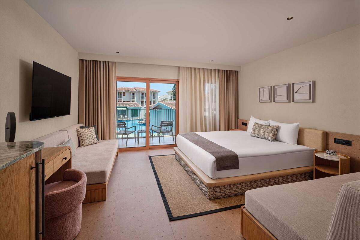 Een hotelkamer met een groot bed, een bureau met stoel, een zithoekje en een balkon met uitzicht op het zwembad.