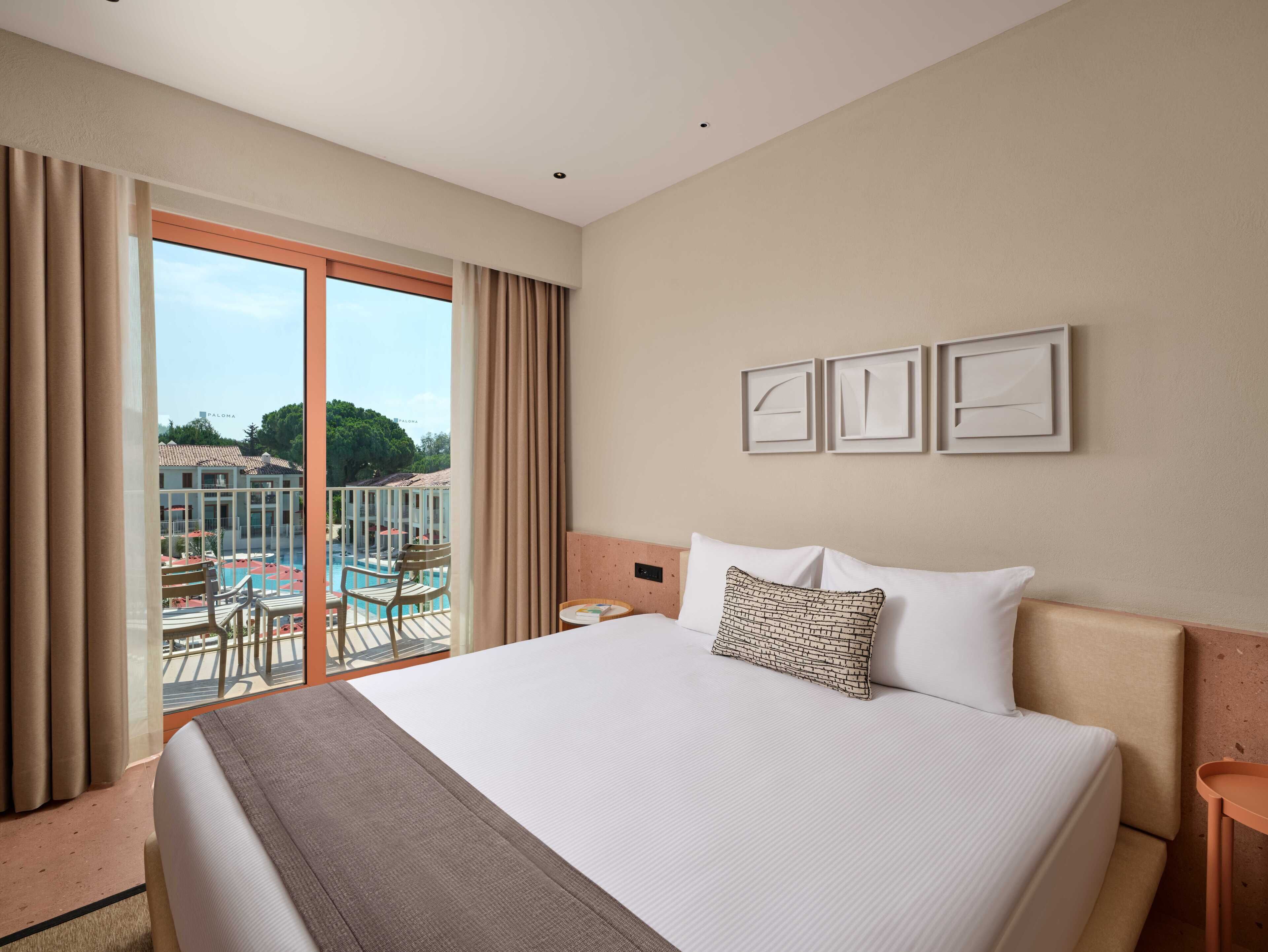 A hotel room with a large bed, a view of a pool and outdoor area, and three framed pictures on the wall.
