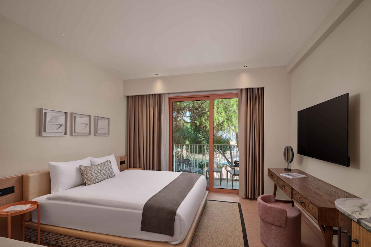 Hotelkamer met een groot bed, een bureau met spiegel, een flatscreen televisie en een balkon met zitplaatsen buiten.