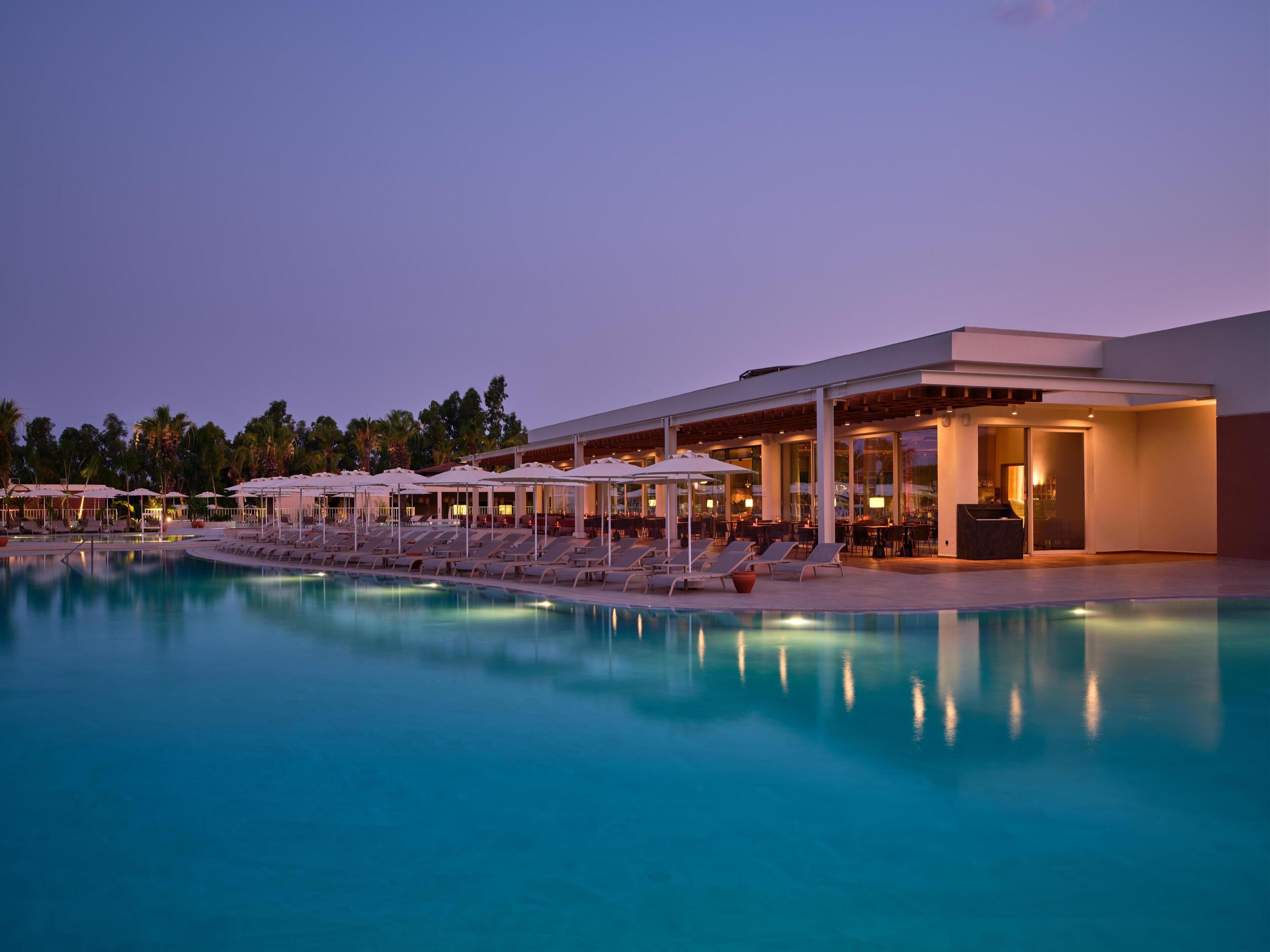 Un espace aménagé au bord de la piscine, avec des chaises longues et des parasols, un bar et un bâtiment doté de grandes fenêtres.
