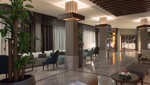 Un vestíbulo amplio con diseño moderno, que incluye una zona de estar con sofás y sillones, una gran planta en maceta y una pared decorativa con patrones geométricos.