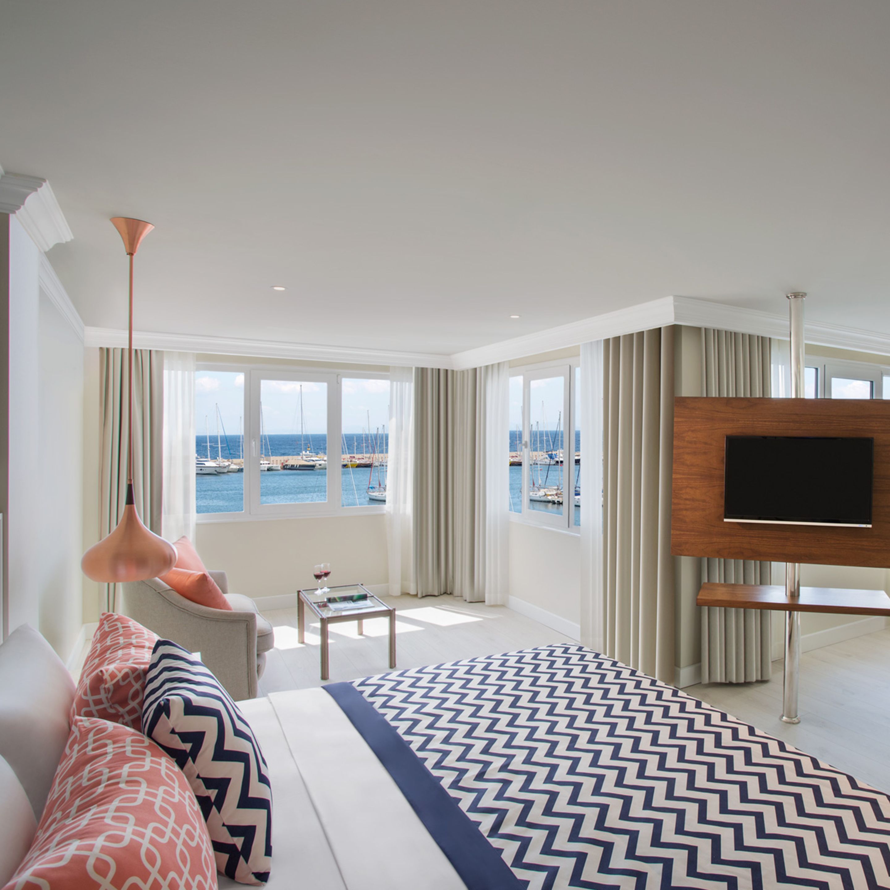 A hotel room with a large bed, a TV mounted on the wall, and a view of the sea through the windows.