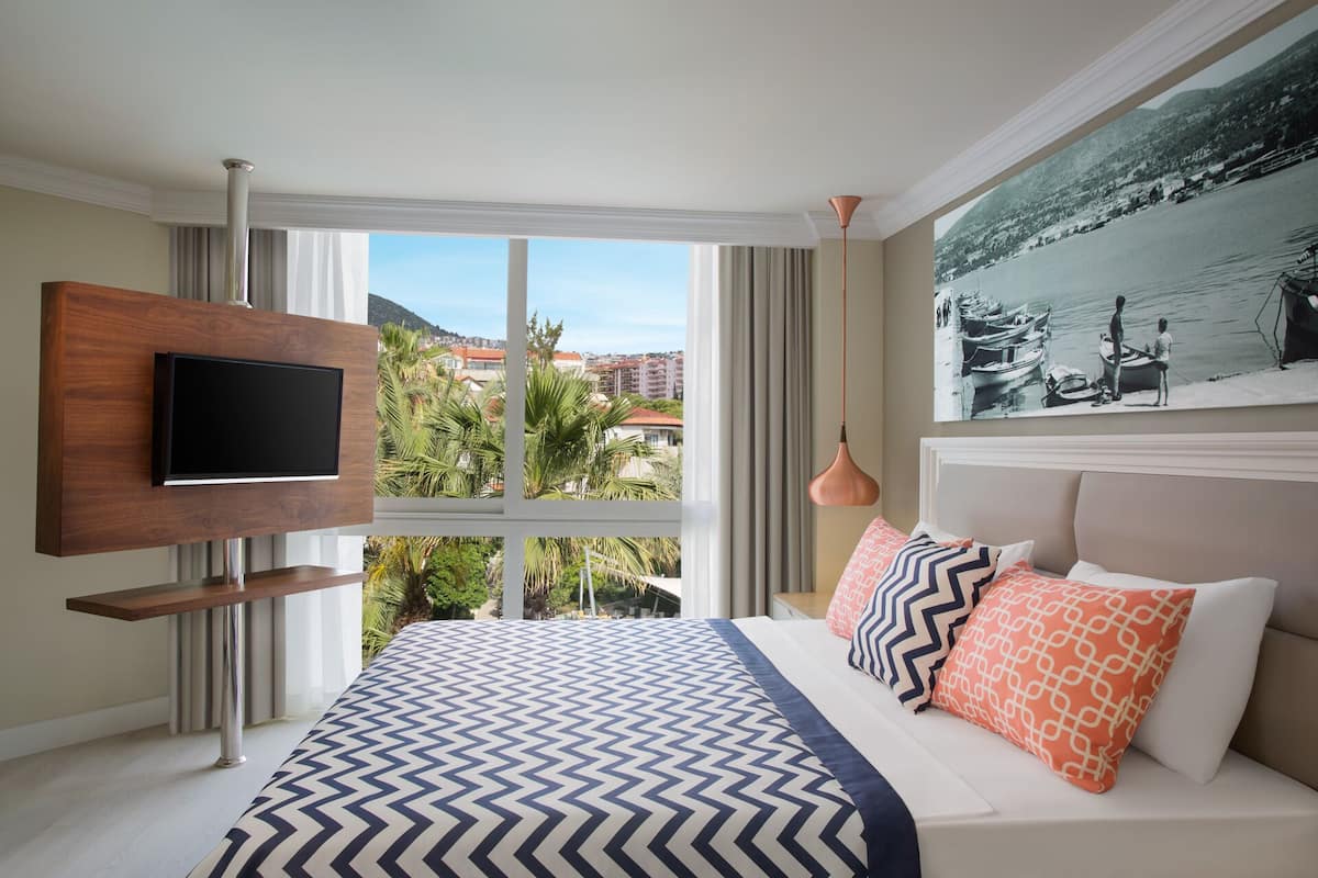 Une chambre d’hôtel avec un lit, un téléviseur fixé au mur, une grande fenêtre donnant sur des immeubles et des arbres, et un tableau accroché au mur.