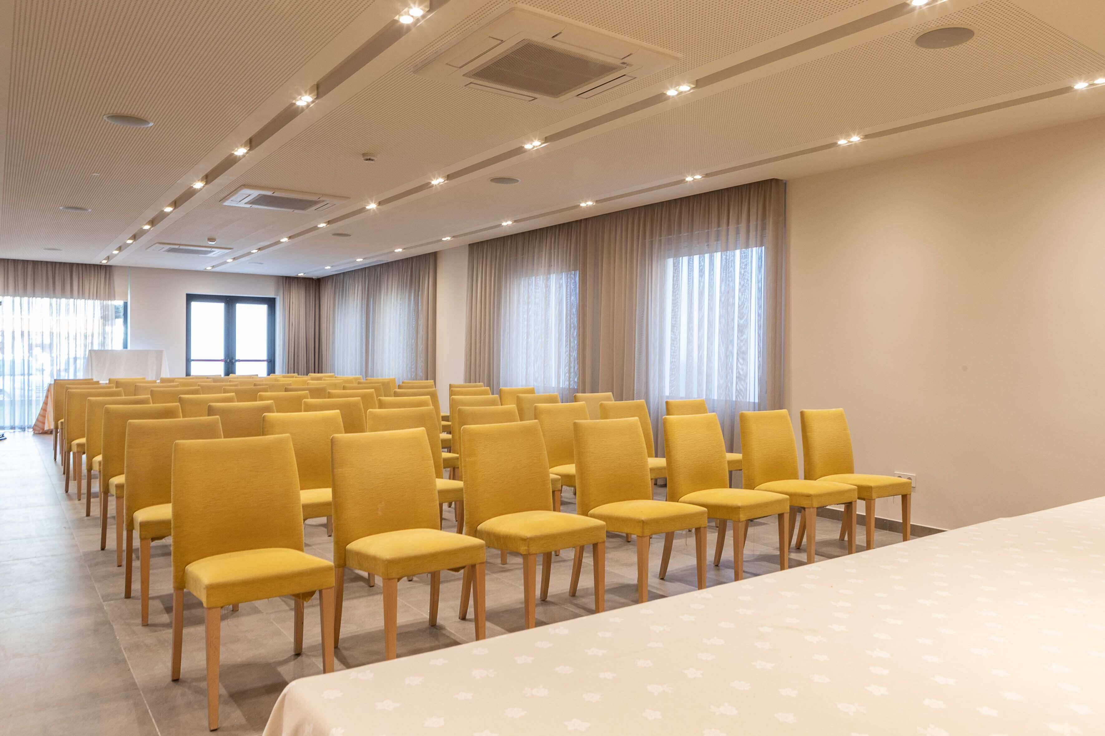 A conference room with rows of yellow chairs and a large table, equipped with ceiling lights and windows with curtains.